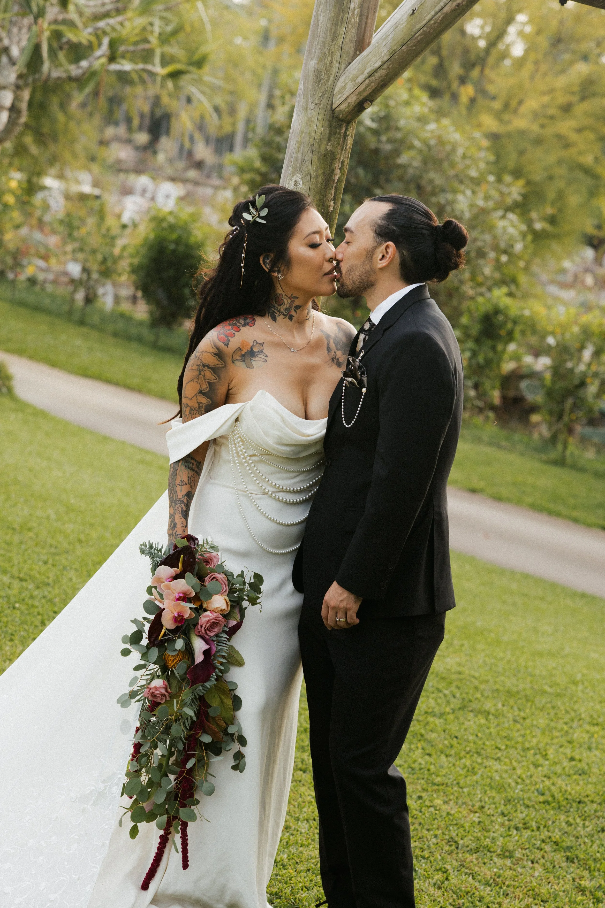 A same-sex couple in wedding attire sharing a kiss outdoors in a park.
