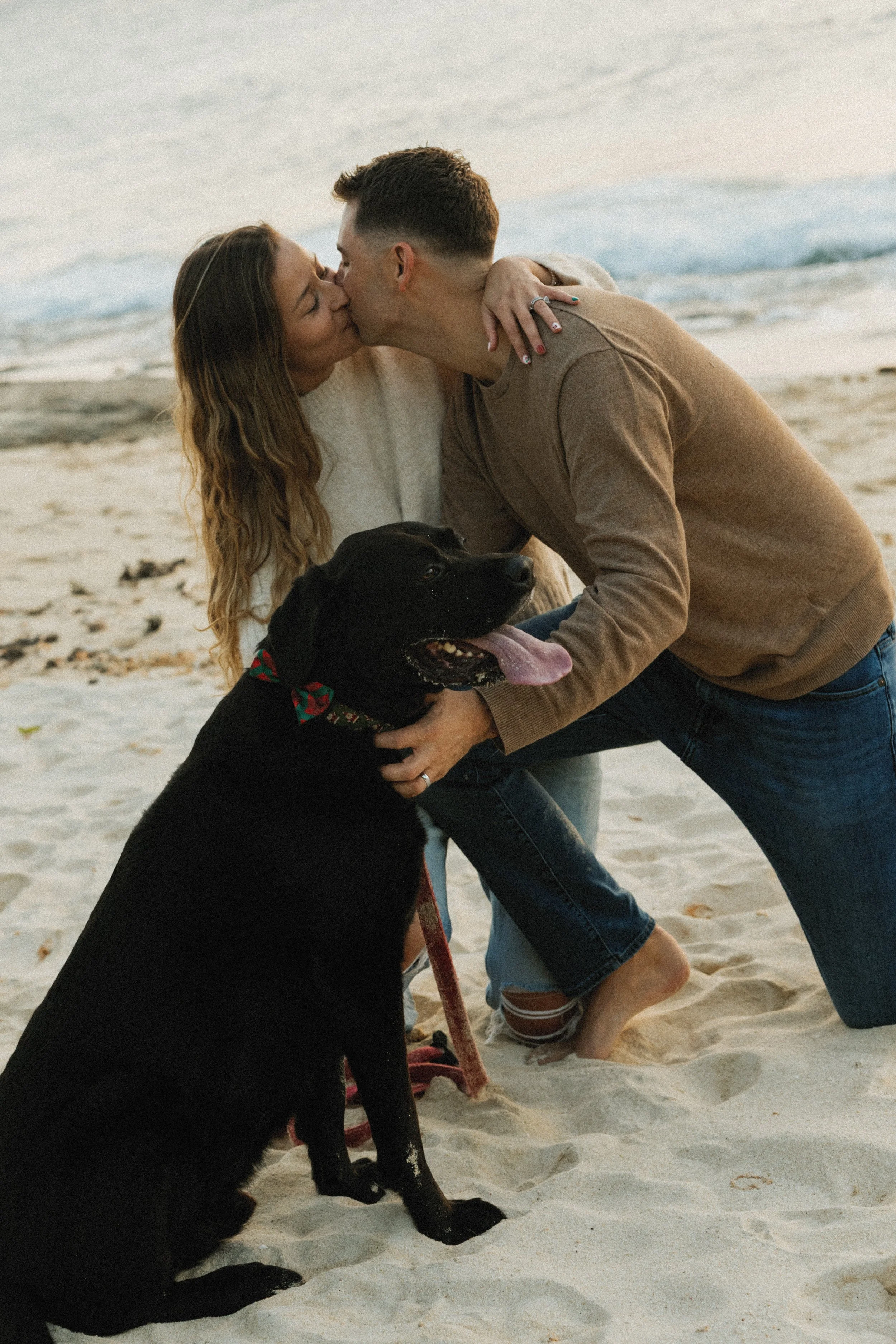 A couple kisses on a beach with a black dog sitting in front of them.