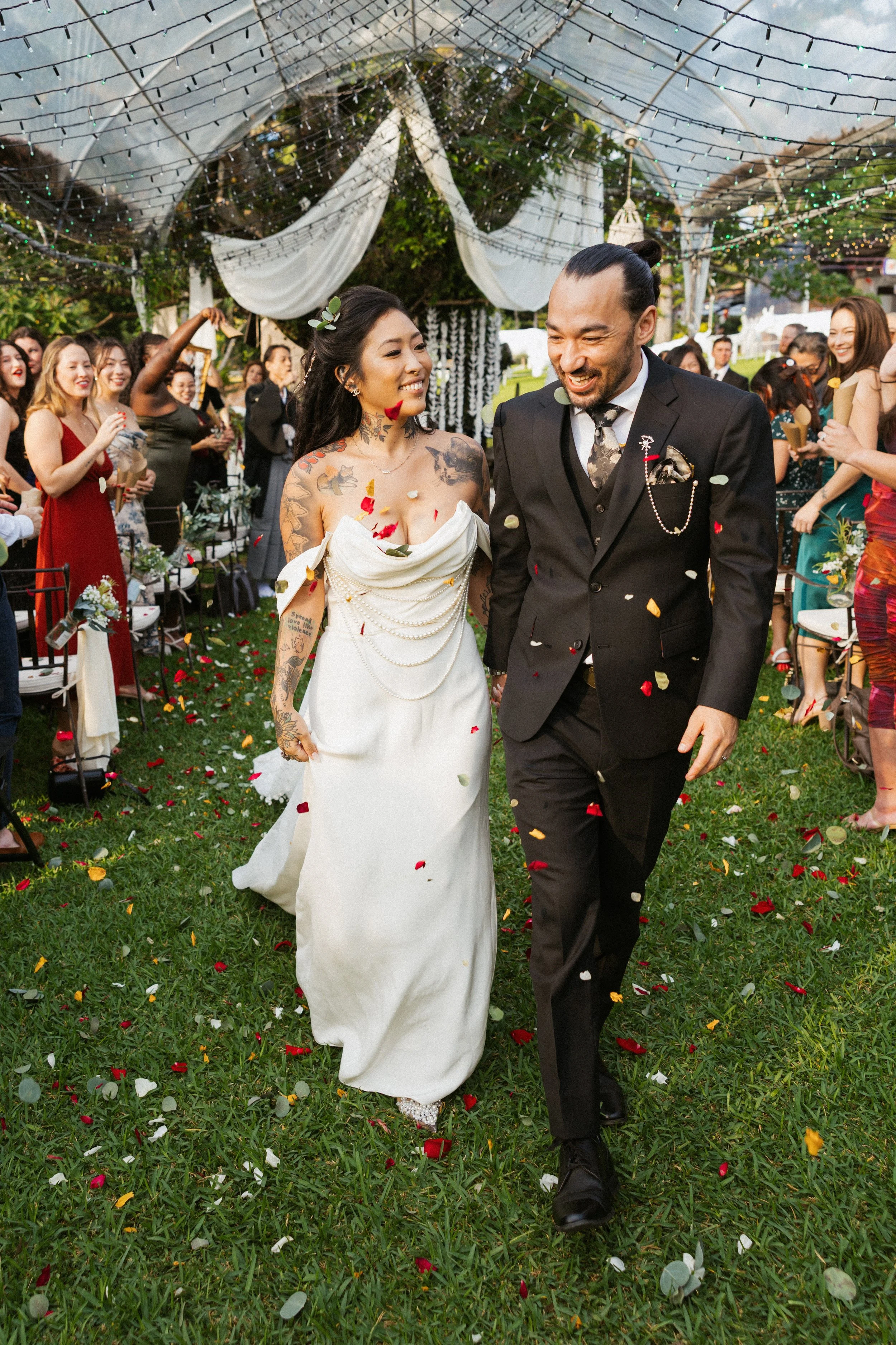 A newlywed couple walking down the aisle at their wedding reception, surrounded by friends and family, with confetti falling around them outdoors under a decorated canopy.