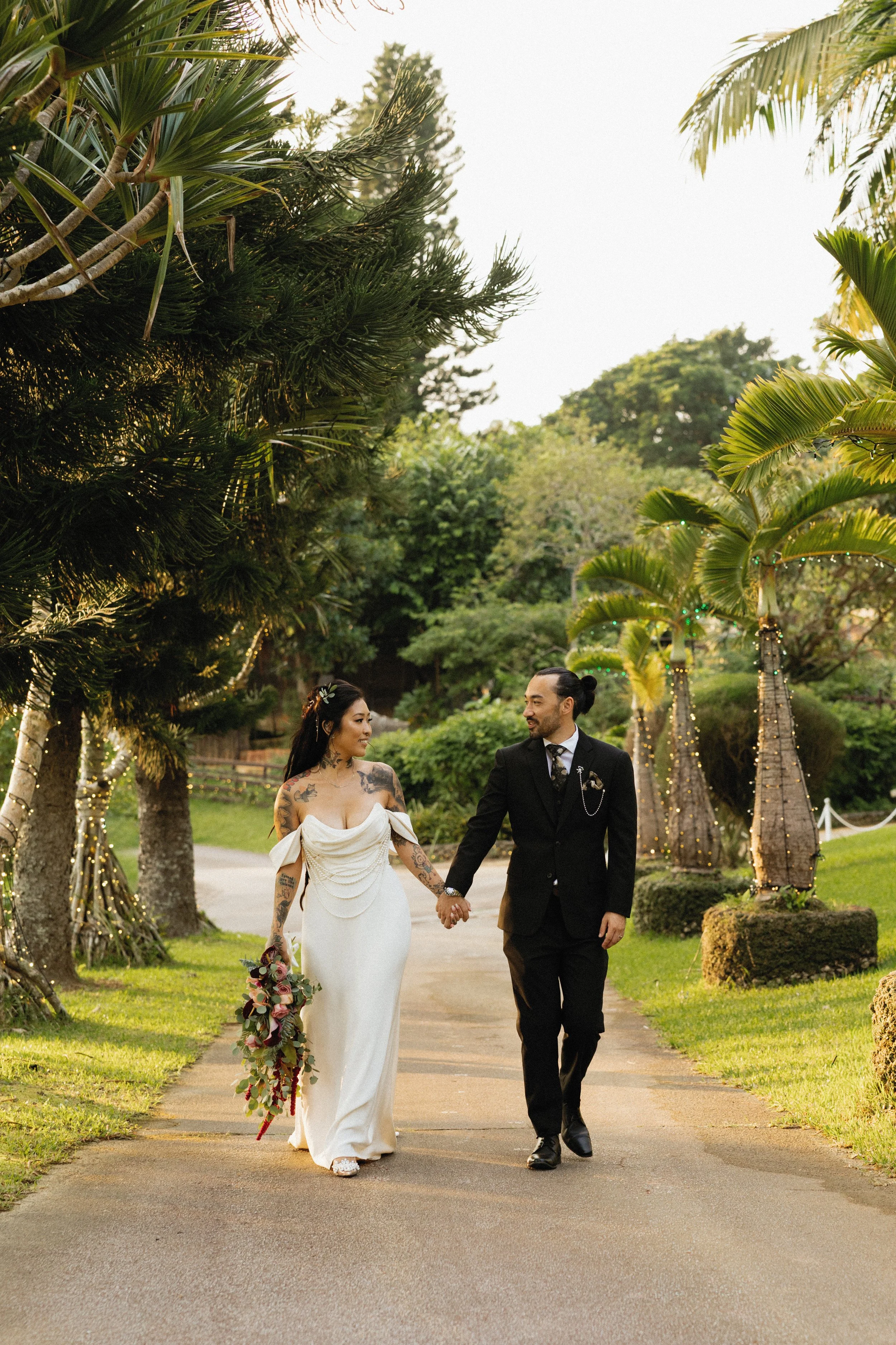A couple in Okinawa dressed in wedding attire holding hands, walking on a pathway in a lush garden with palm trees and fairy lights.