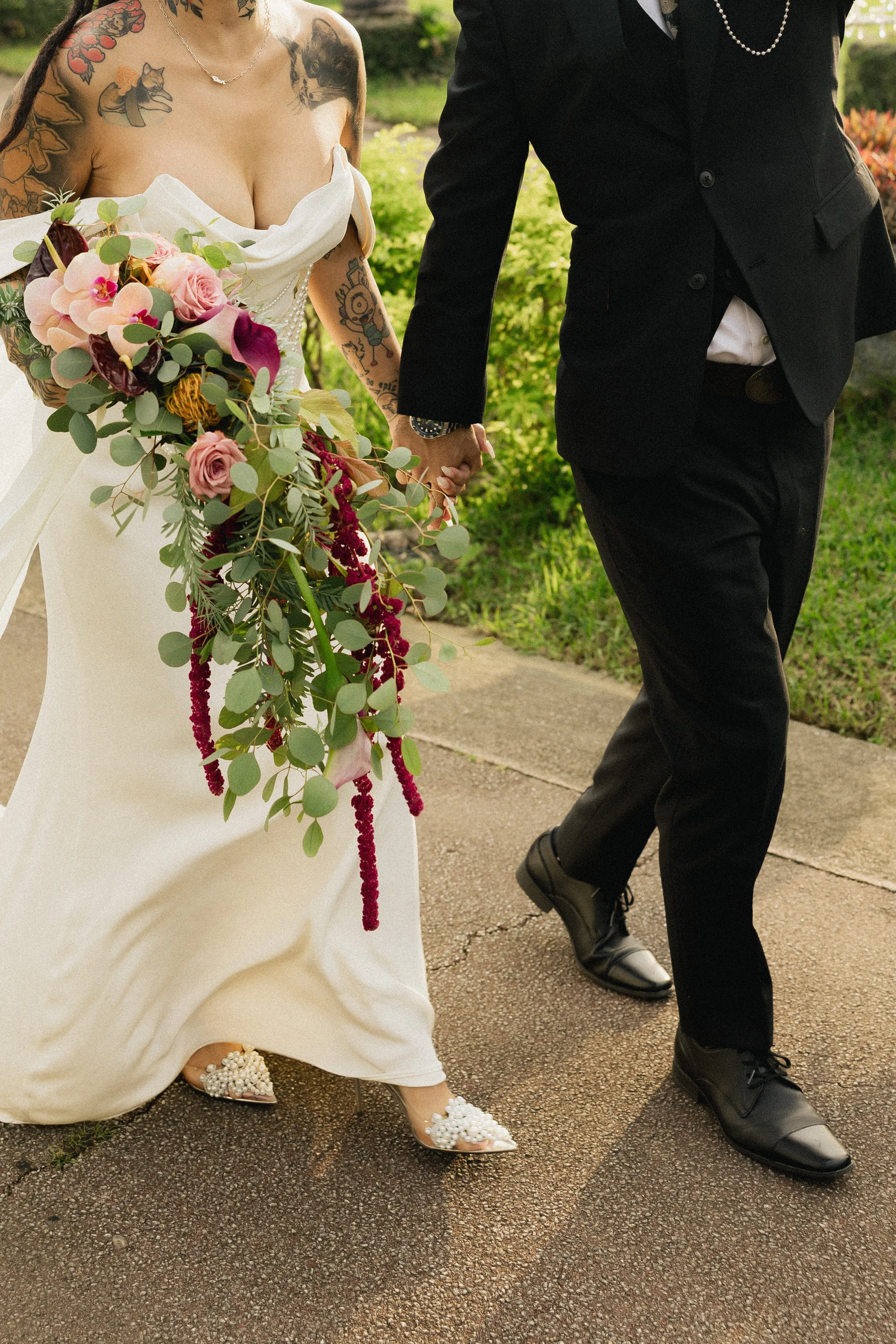 A bride and groom holding hands and walking outdoors on a wedding day. The bride is in a white dress with floral embellishments on her shoes, and she is holding a large bouquet of pink, red, and purple flowers and greenery. The groom is in a black su