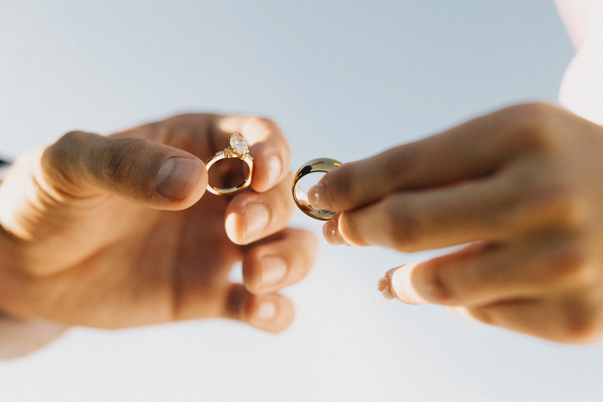 Close-up of a person's hands holding two gold rings, one with a diamond, against a clear sky.