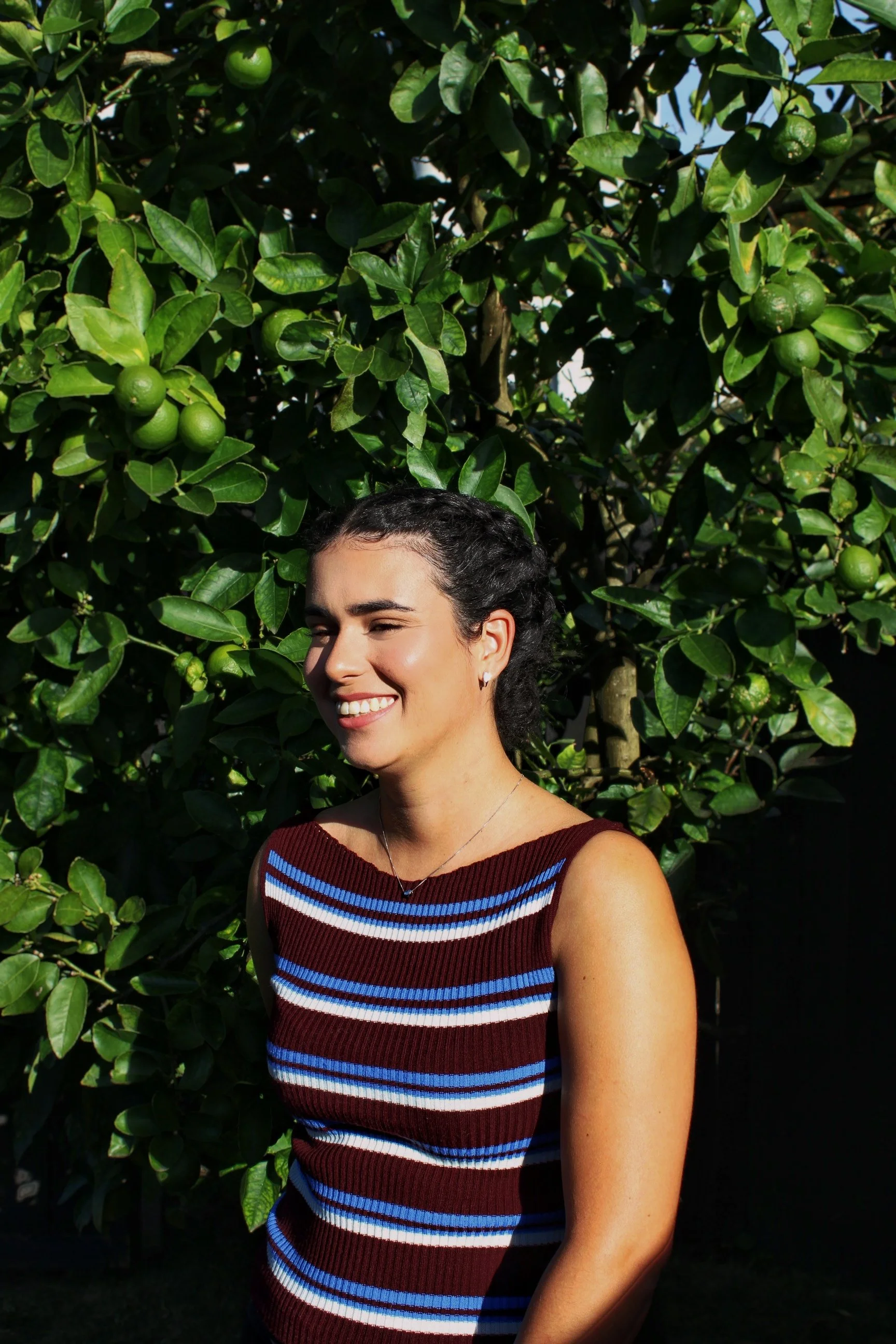 A woman with dark hair pulled back, smiling, standing outdoors in front of a lemon tree with green lemons and leaves.
