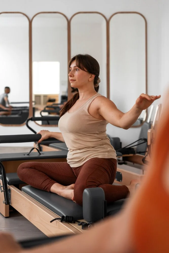 Une femme en tenue de sport effectue un exercice d'étirement ou de méditation en position de tailleur sur un équipement de pilates dans une salle de sport. Un miroir et d'autres personnes s'entraînant sont visibles en arrière-plan.
