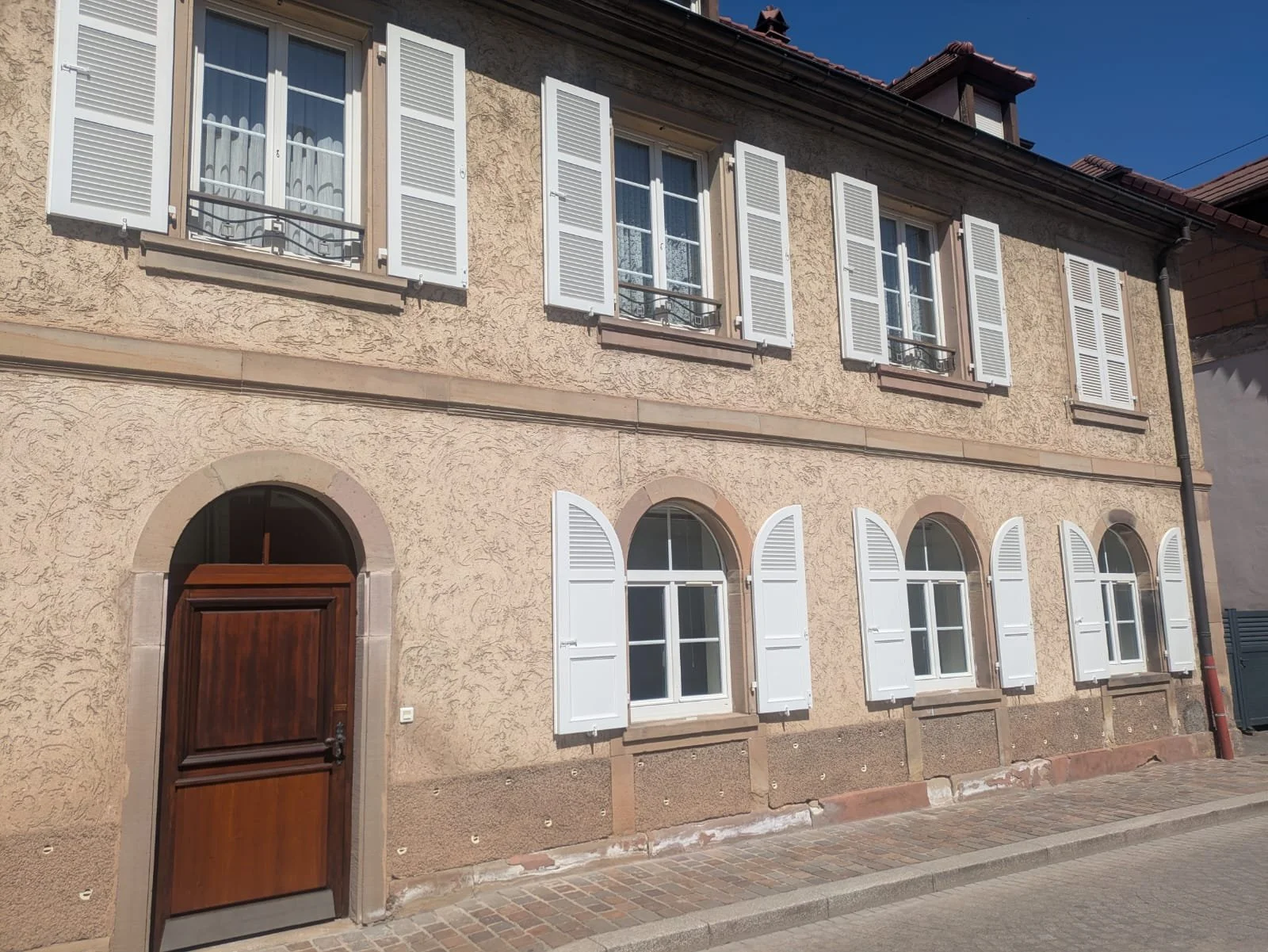 Façade du studio Pilates "La parenthèse" à Molsheim avec des fenêtres à volets blancs, porte en bois foncé, murs en stuc beige, sous un ciel bleu clair.