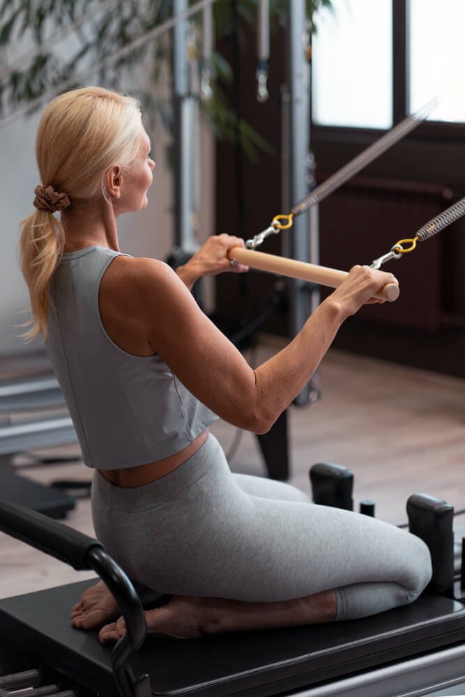 Une femme blonde pratique des exercices de Pilates avec un appareil à la maison, assise à genoux sur un tapis, tirant sur des cordes de résistance.