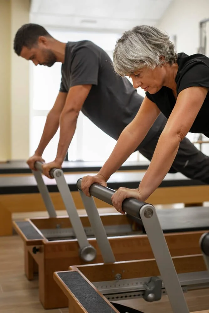 Deux personnes, un homme et une femme, pratiquent le Pilates sur des reformers dans une salle de gym. Ils sont penchés en avant, en train de faire un exercice de renforcement musculaire.