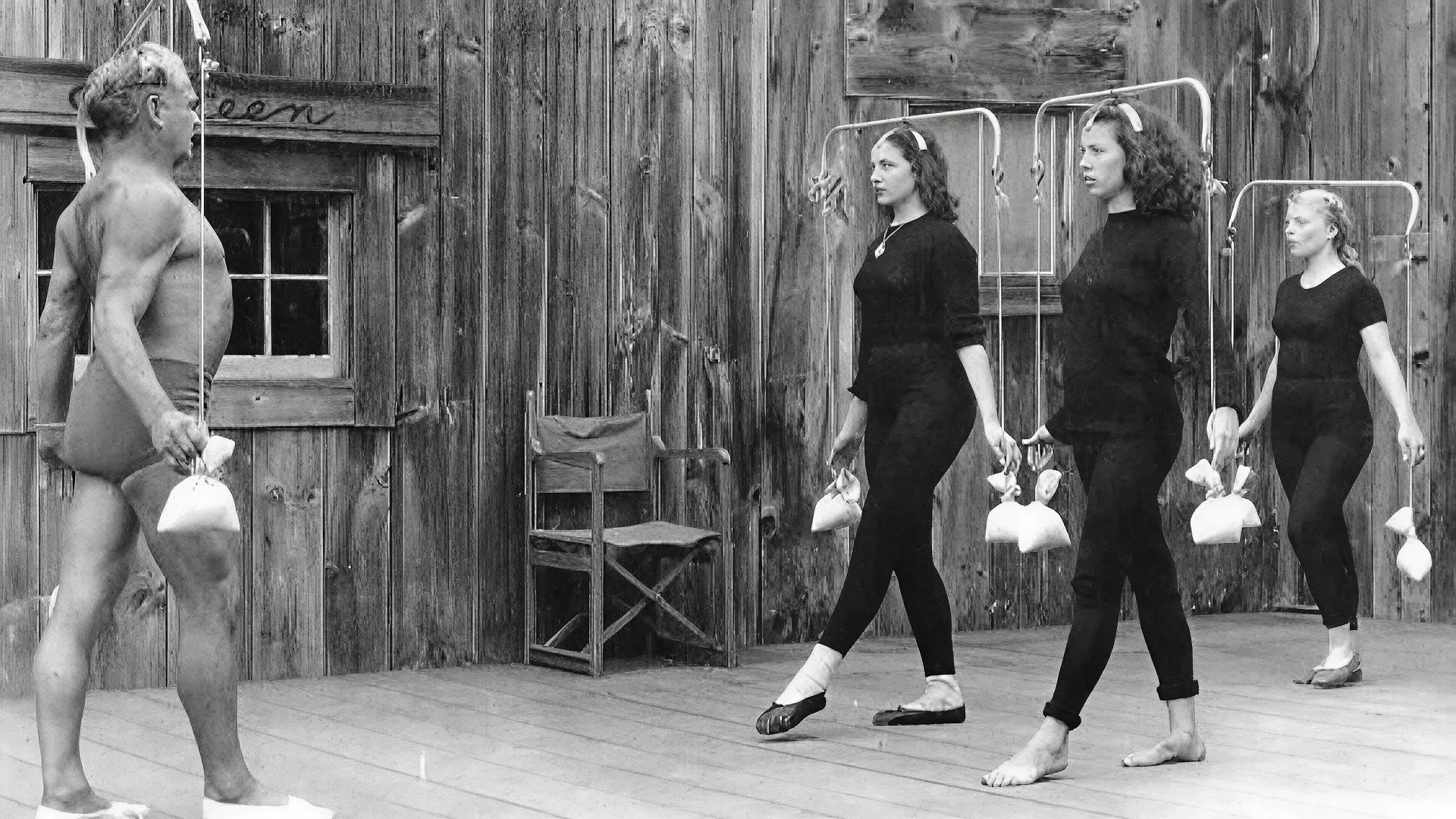 Une scène en bois avec quatre personnes, dont un homme nu avec des sacs en plastique, et trois jeunes femmes habillées en noir, portant des sacs en plastique, dans une posture de ballet.