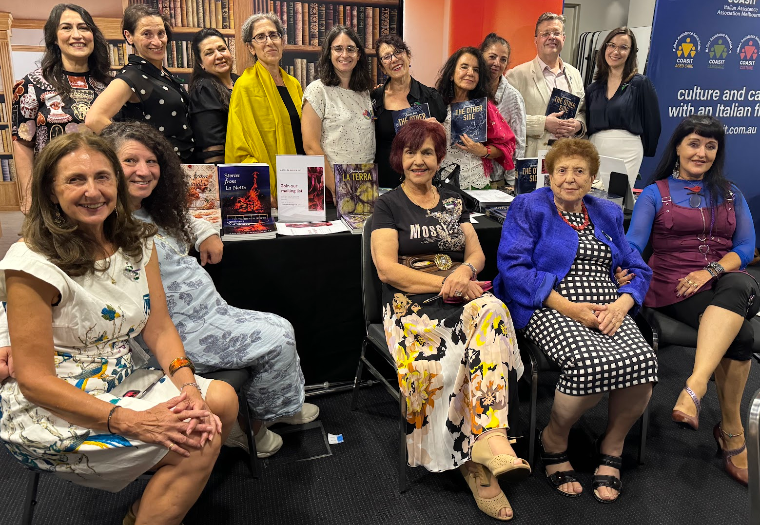 A group of women and one man gathered at a book signing or literary event, with books displayed on a table and a blue banner with logos and text in the background.