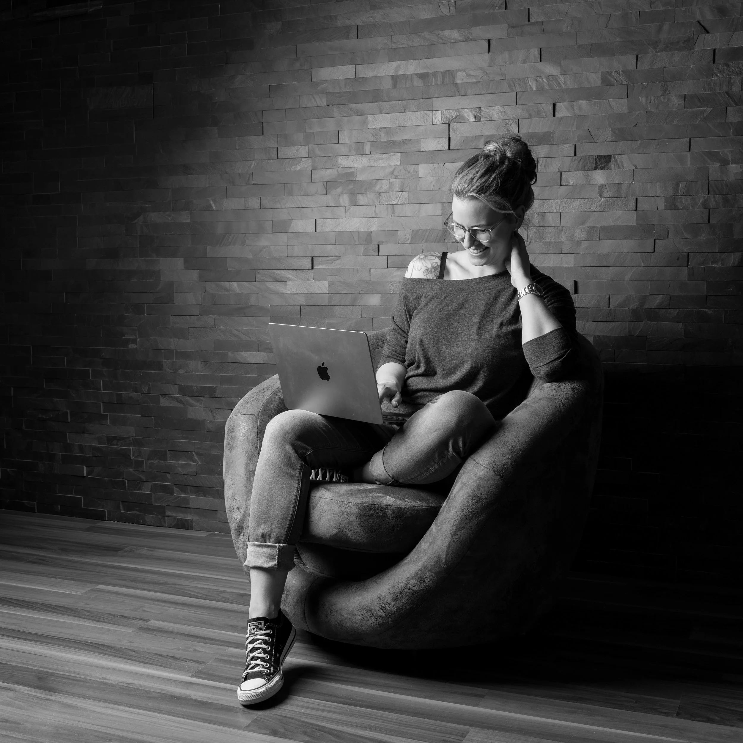 A woman with glasses and a bun hairstyle sitting on a curved chair against a textured wall, laughing and looking down at her laptop, wearing a sweater and sneakers.