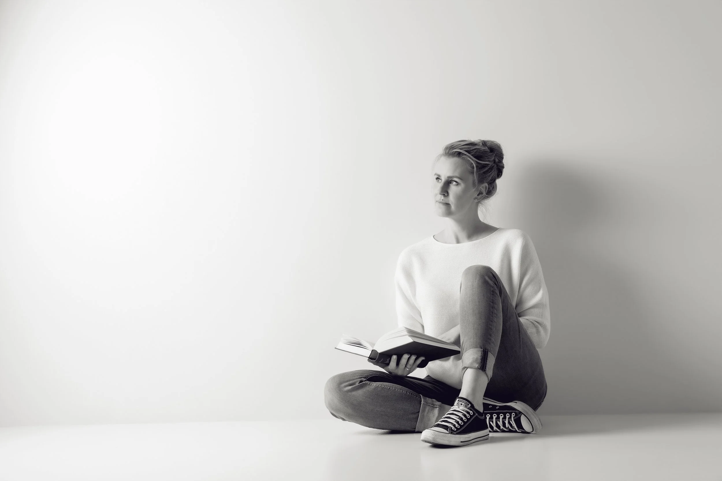 Black and white photo of a woman sitting cross-legged on the floor, holding a book, wearing casual clothes and Converse sneakers, against a plain wall.