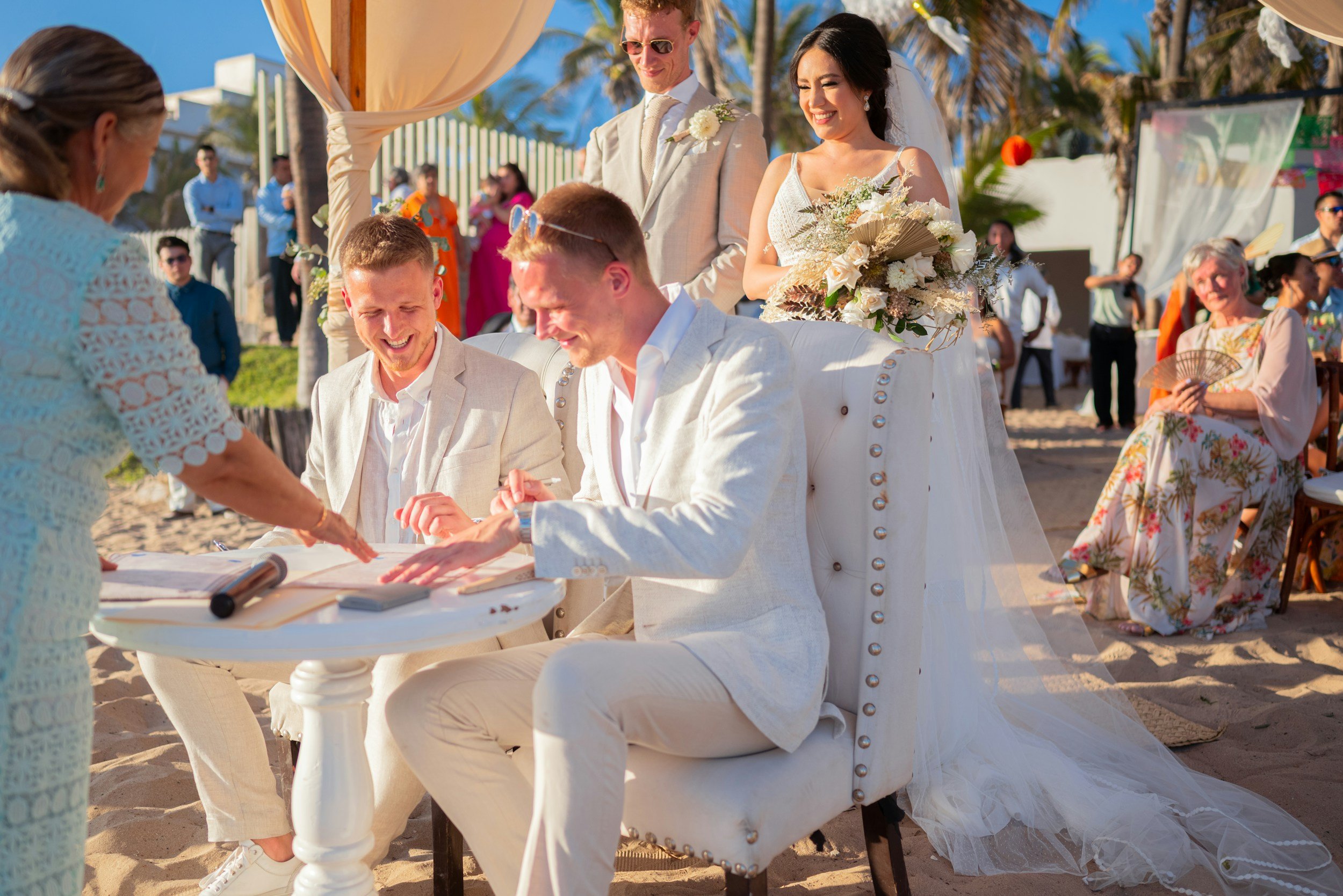 A wedding ceremony on the beach with the bride and groom kneeling and signing documents, surrounded by guests, under a sunny sky with palm trees.