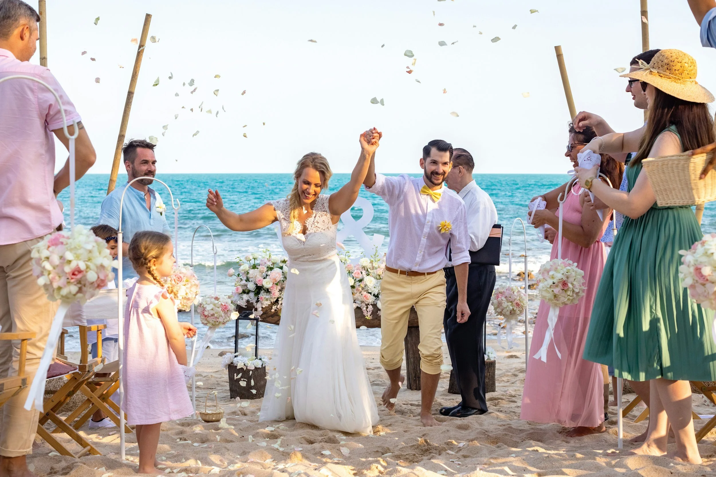 A wedding ceremony on the beach with the bride and groom walking hand in hand, surrounded by guests, floral arrangements, and ocean in the background, as confetti falls from above.