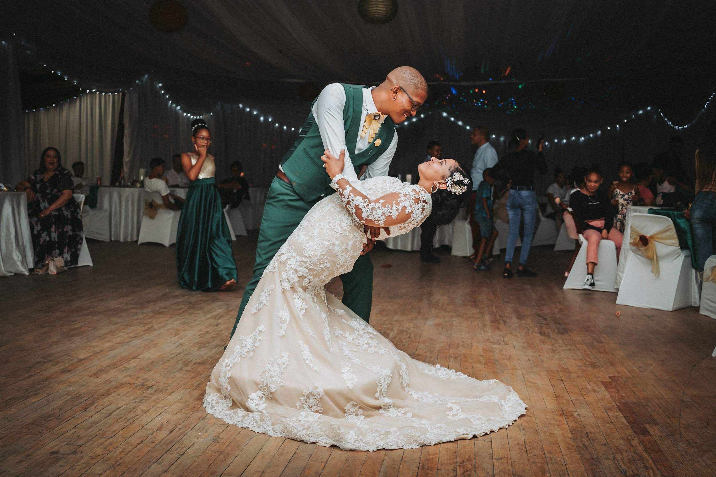 A bride and groom are dancing together at a wedding reception in a decorated venue with string lights. The groom is leaning in toward the bride, who is smiling and looking up at him, wearing a lace wedding gown. Guests are seated and standing in the background, watching and enjoying the moment.