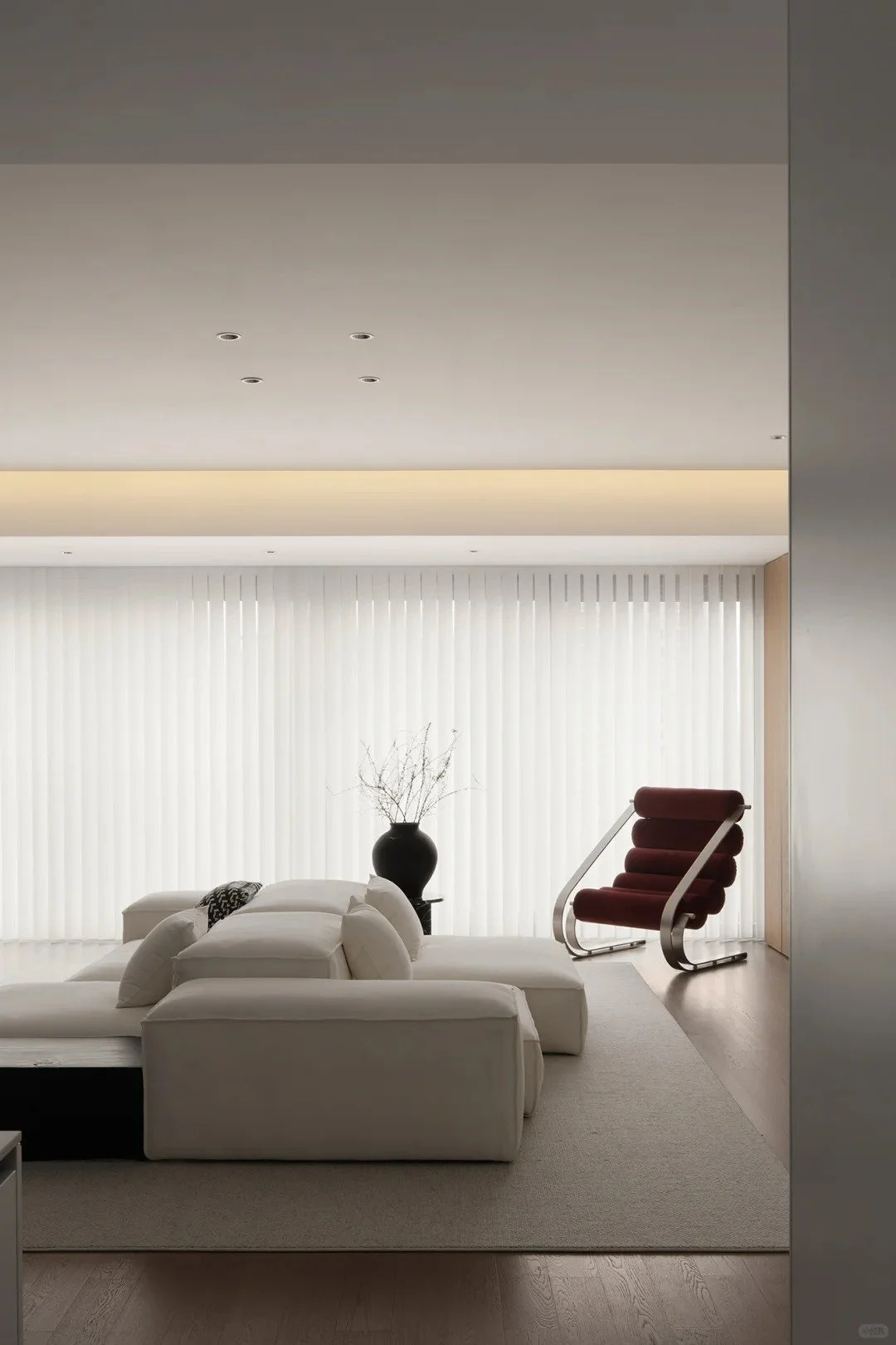 Modern living room with white sectional sofa, black vase with decorative branches, and a red lounge chair near window with vertical blinds.
