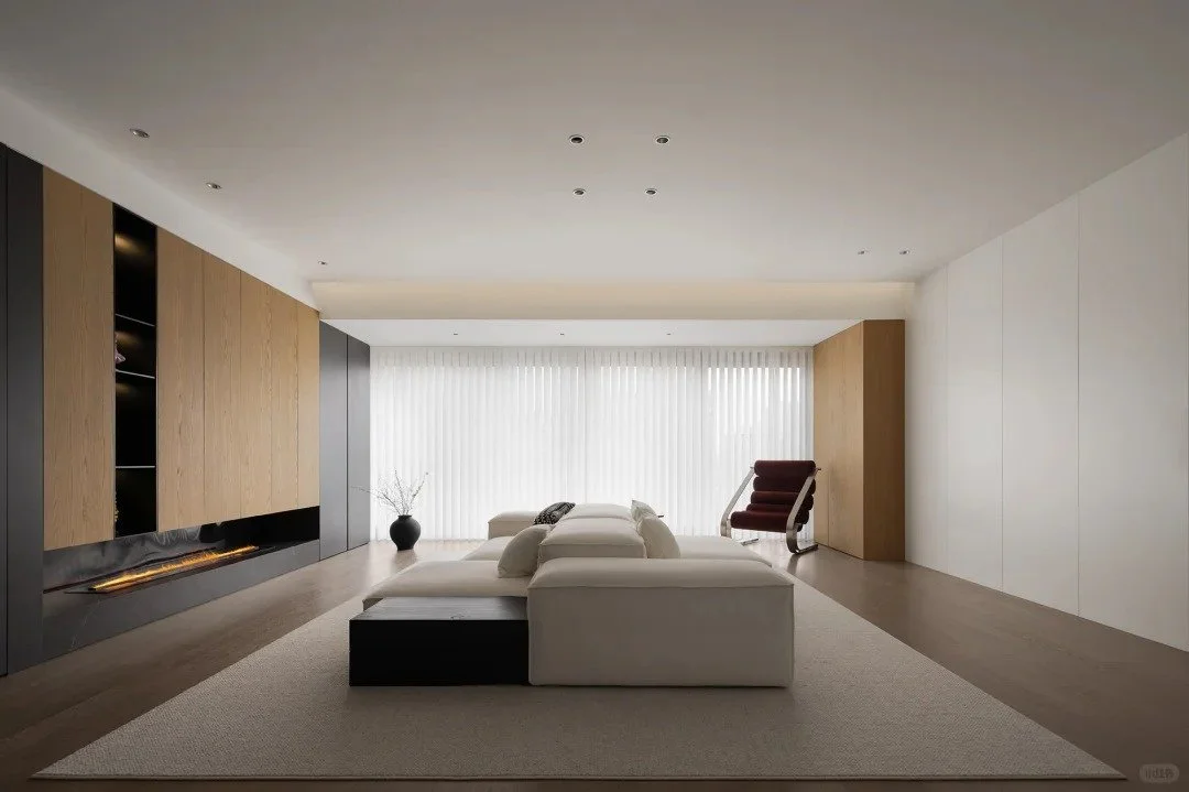 Modern living room with white sectional sofa, black side table, brown accent chair, large window with vertical blinds, wooden and white wall panels, and decorative vase with branches.