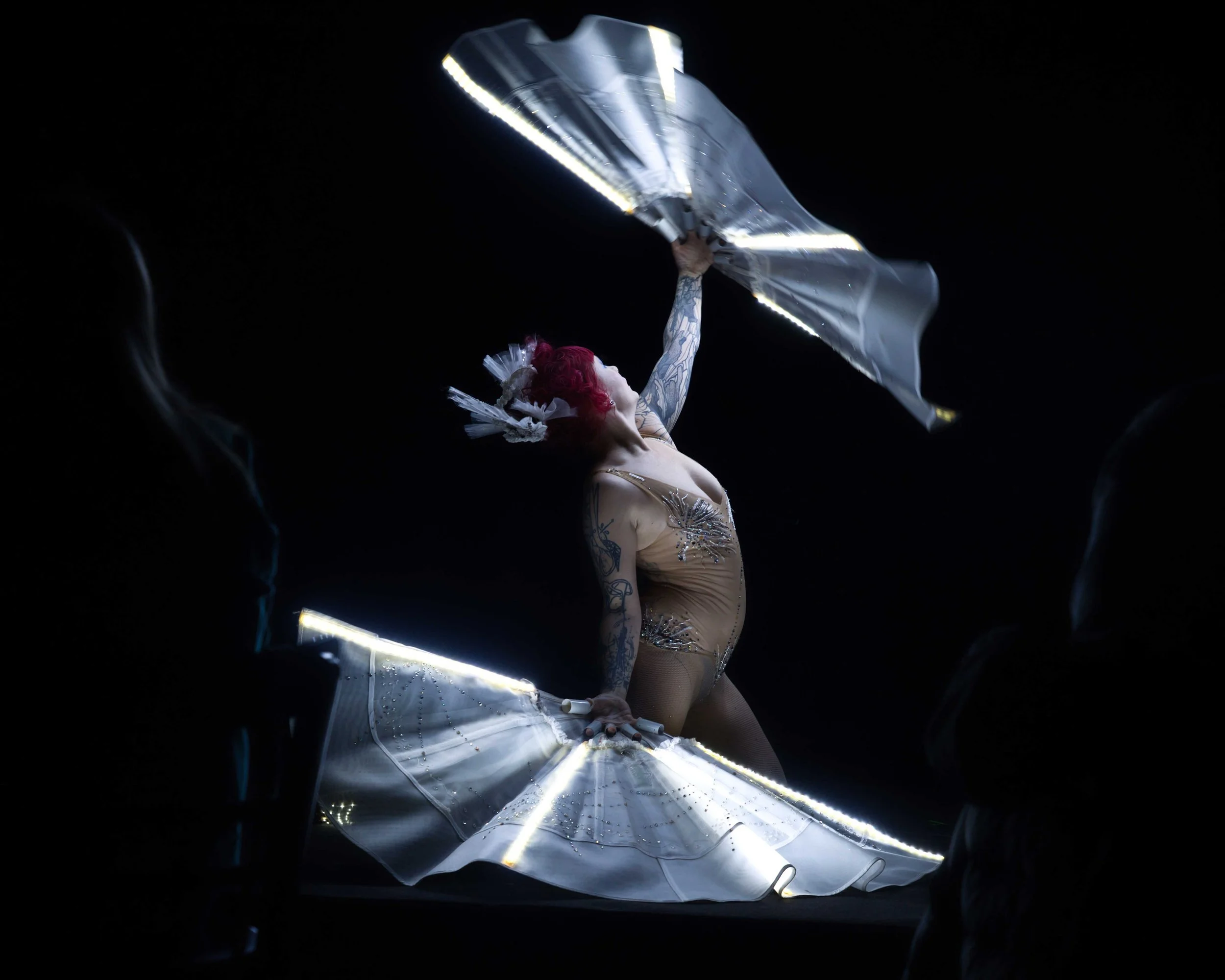 Eine Stelenschlange mit rosa Haaren, die tanzend einen reflektierenden Schirm hebt, umgeben von dunklem Hintergrund und Silhouetten im Vordergrund.