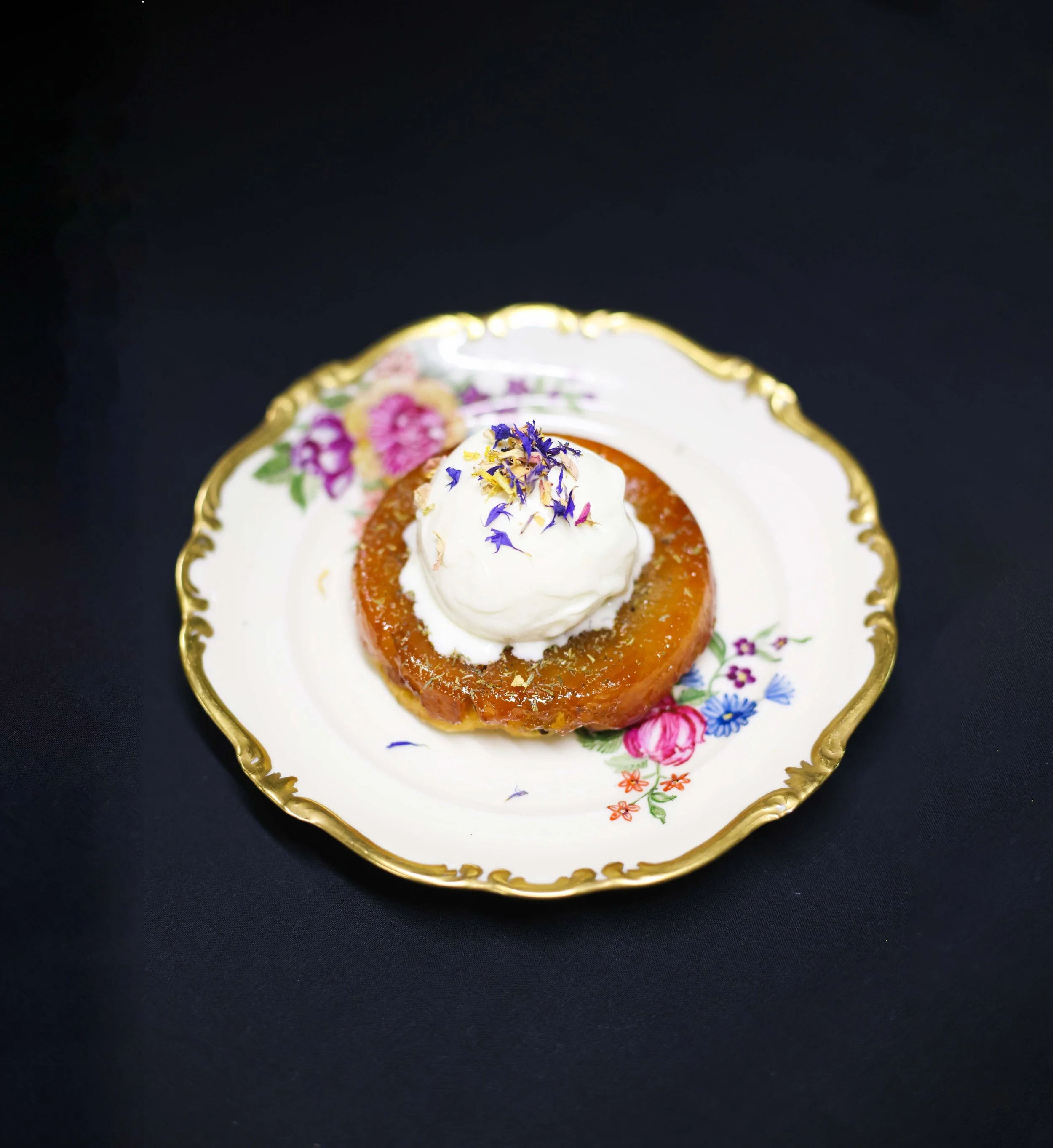 Karamellierter Apfel mit Sahne und Blumenblüten auf einem eleganten Porzellan-Teller mit goldener Kante auf schwarzem Hintergrund.