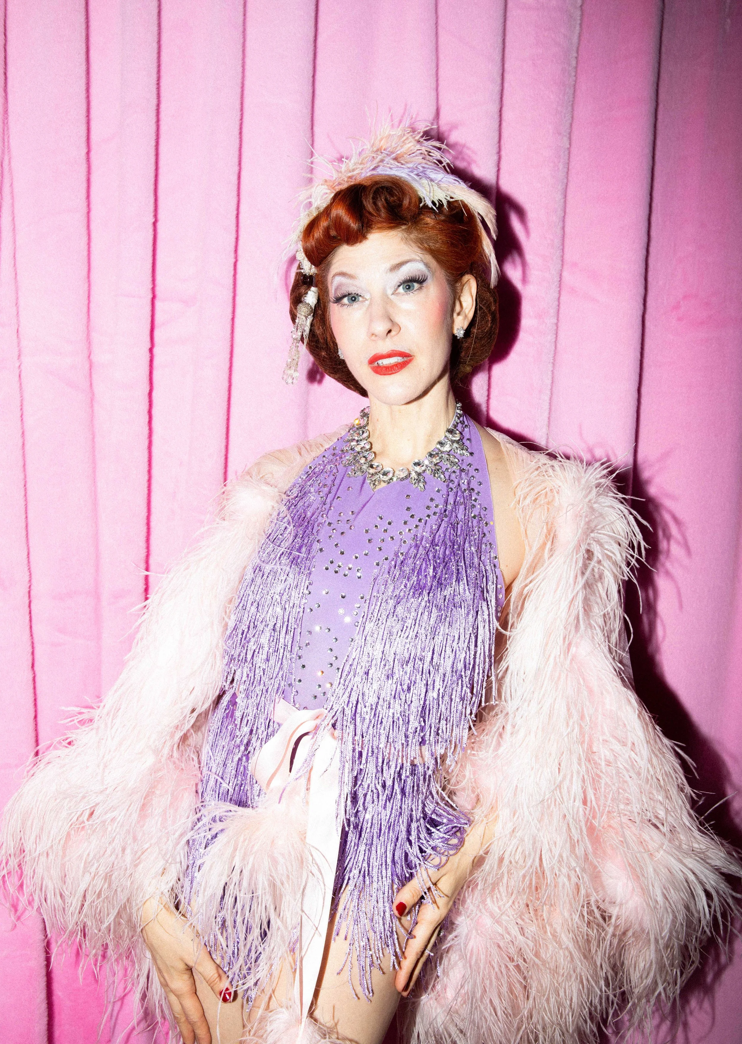 Frau in 1950er Jahre Mode, trägt einen lilafarbenen Fransenrock, einen Pelzmantel und ein Schmuckset, steht vor pinkem Vorhang.