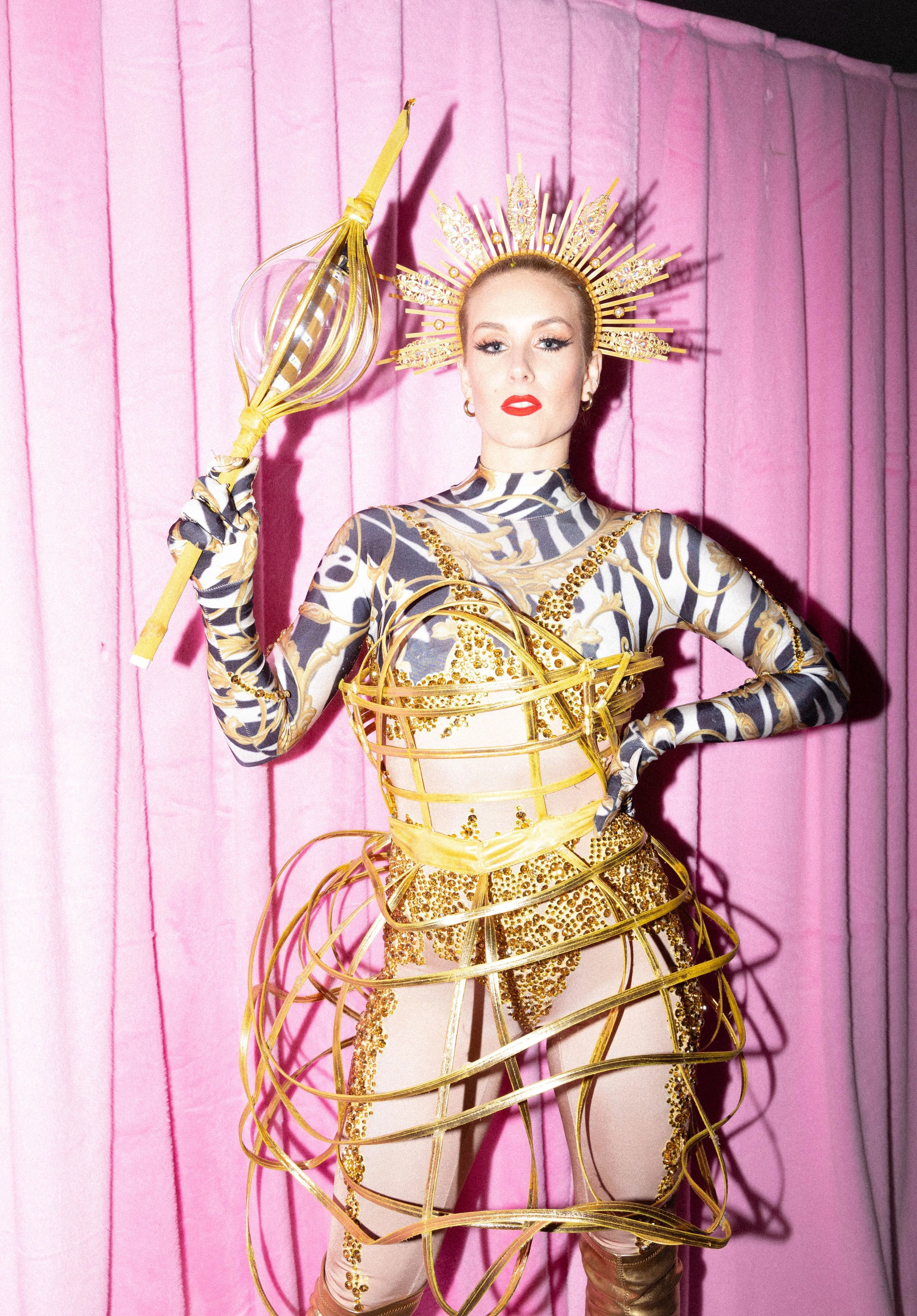 Frau in auffälligem Kostüm mit goldenen Ringen und Kopfkrone vor pinkem Hintergrund.