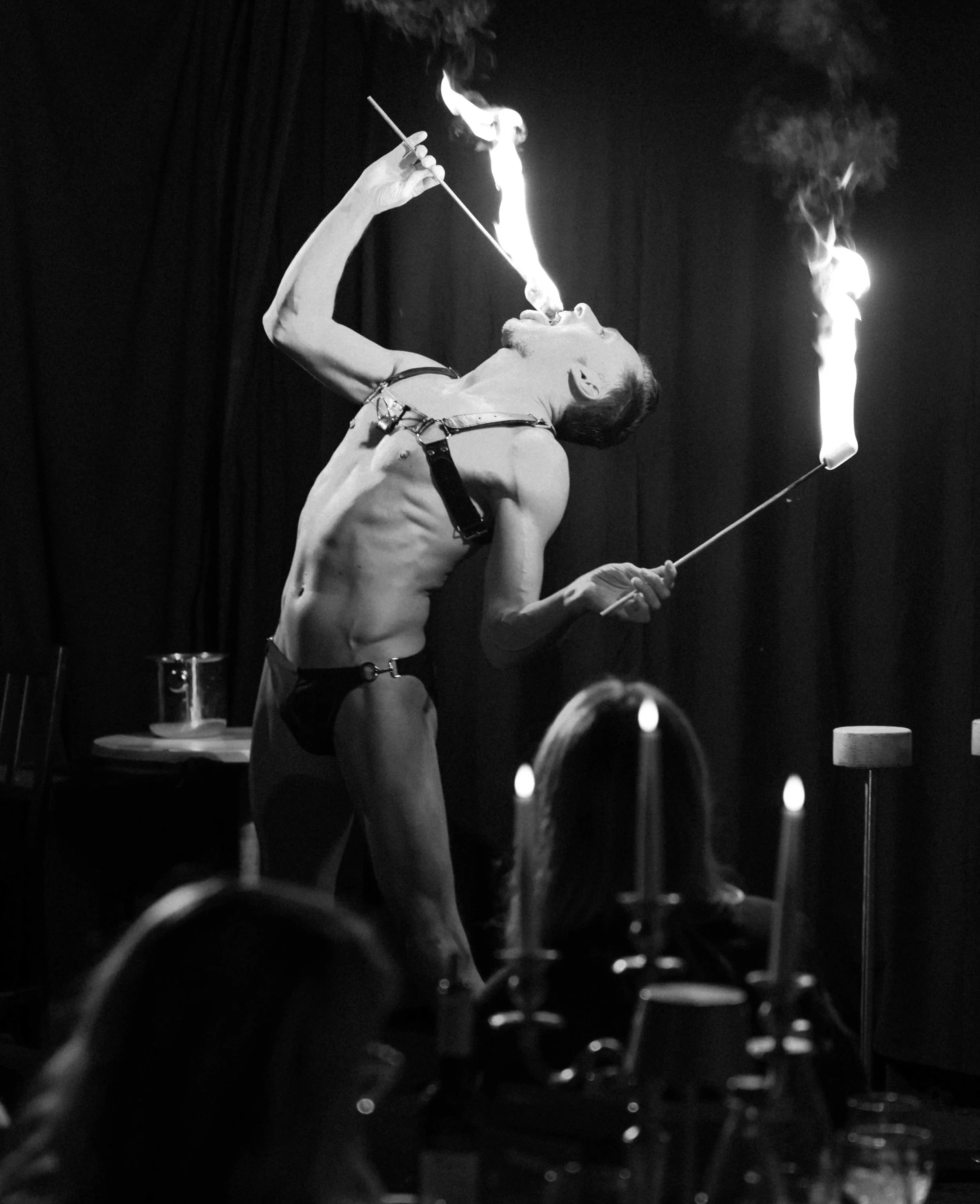 Ein nackter Mann beim Feuerspucken in einem Schwarz-Weiß-Foto, mit dunklem Hintergrund und Kerzen im Vordergrund.