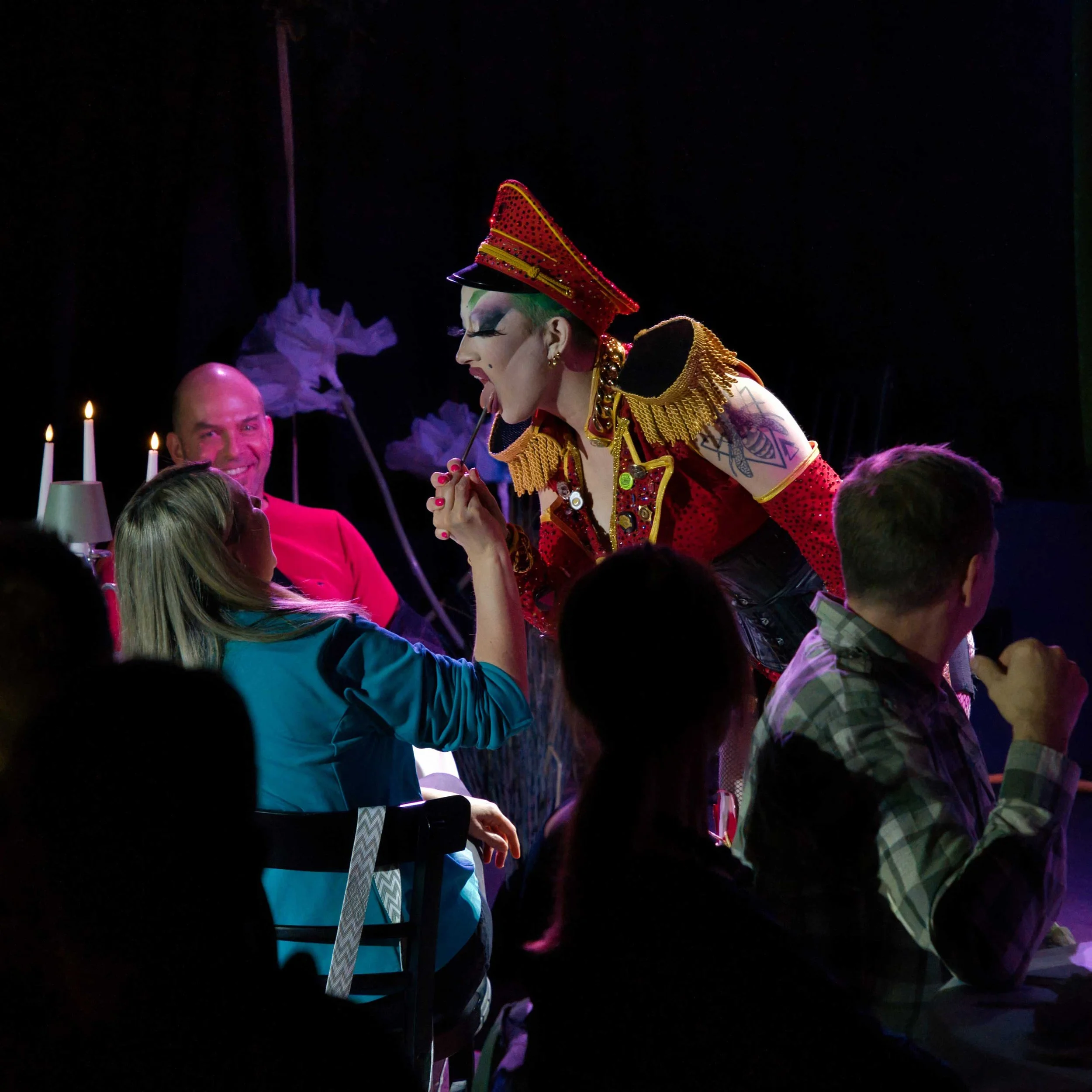 Ein Mann in pinkem Hemd sitzt lächelnd an einem Tisch, während eine Drag Queen in einem roten Militär-Outfit eine Frau am Tisch mit einem Zahnstocher im Mund anspricht. Weitere Personen sind im Hintergrund.