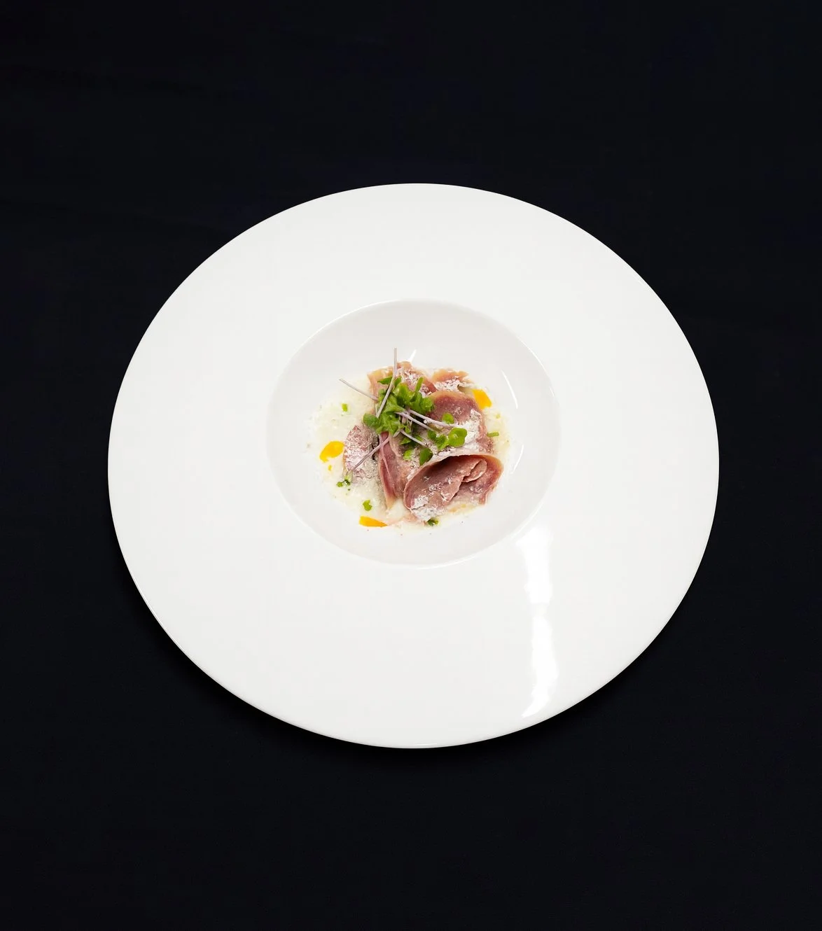 Fein angerichtener Teller mit rohem Fisch, garnierte mit frischen Kräutern und Mikrogreens, serviert auf einem weißen Teller auf schwarzem Hintergrund.