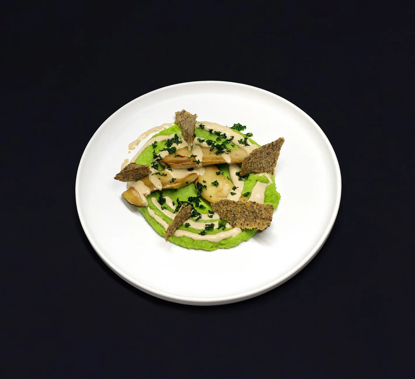 Vorgekochtes Gericht mit grünen und weißen Soßen, geballtem Gemüse, dunklen Brocken und kleinen grünen Kräutern auf weißem Teller