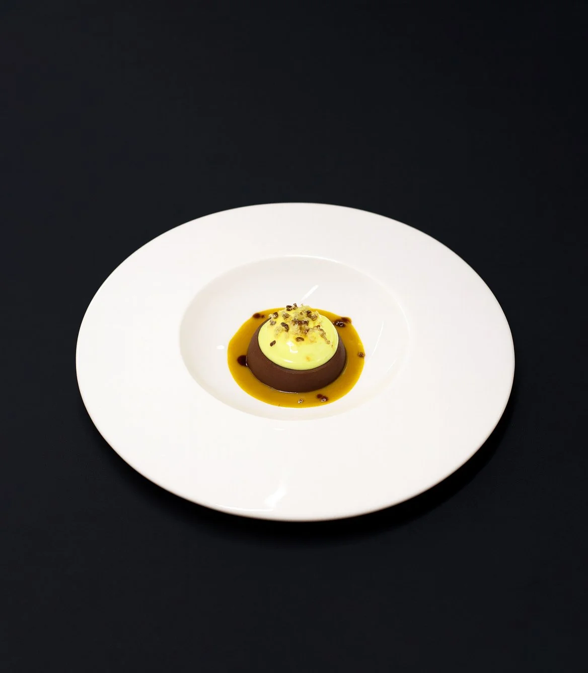 Dessert mit Schokoladen-Mousse, Vanillecreme, Karamellsoße und kleinen Schokostreuseln auf einem weißen Teller vor schwarzem Hintergrund.