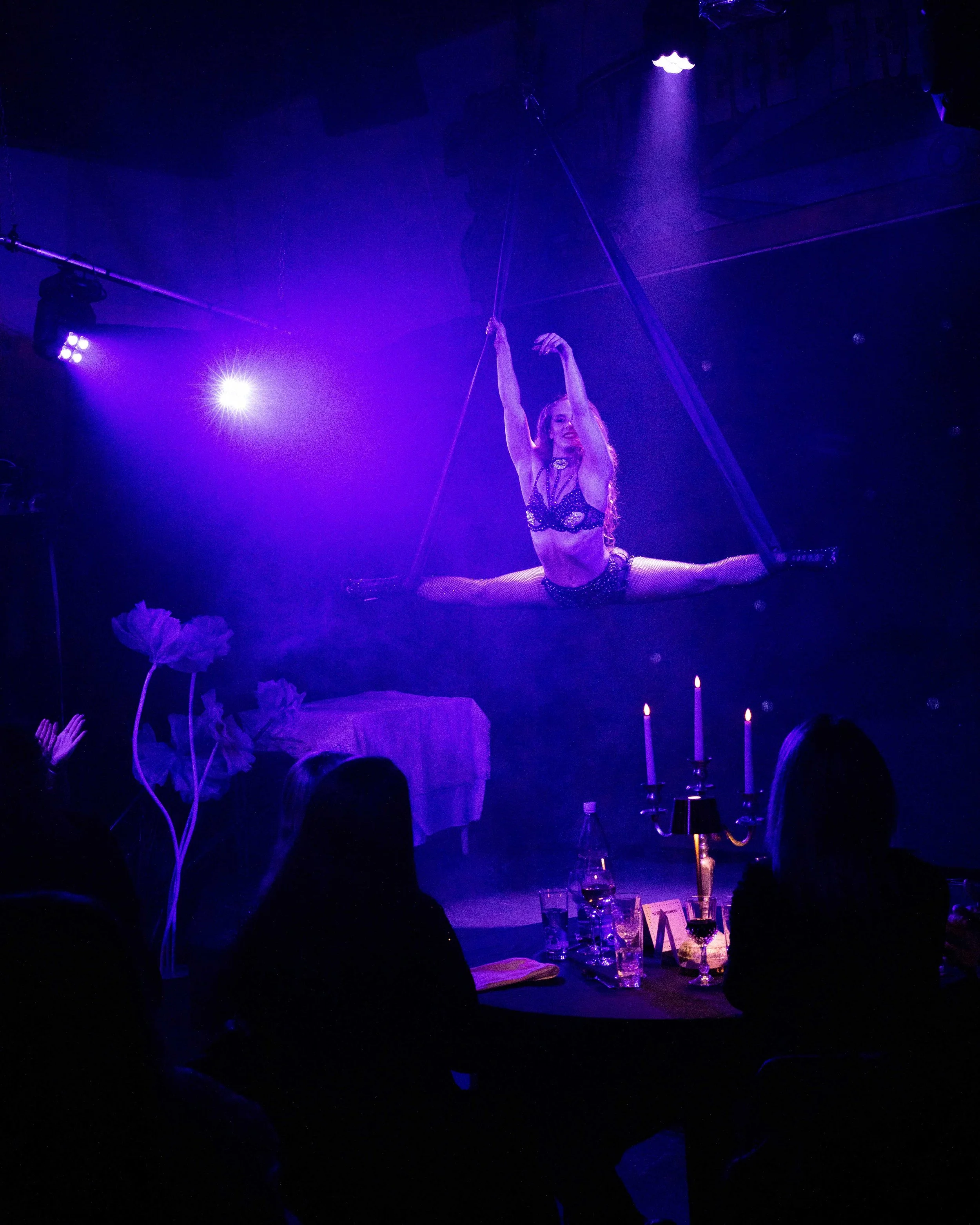Eine Artistin präsentiert eine akrobatische Show auf einer hängenden Trapez, beleuchtet von blauen und violetten Lichtern in einem dunklen Theaterraum.