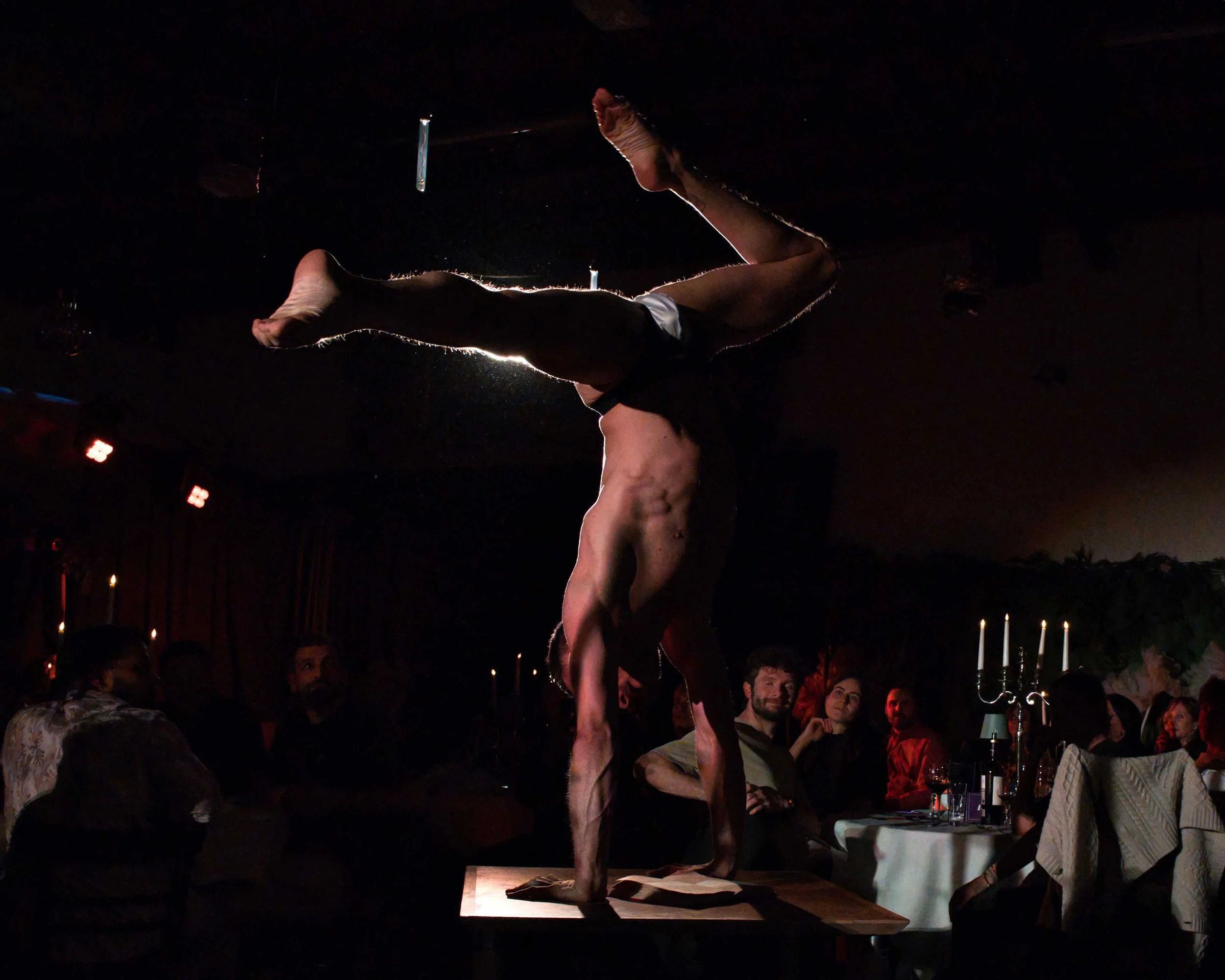 Ein Artist macht eine akrobatische Handstand-Nummer auf einer Tafel vor einem dunklen Publikum in einem gemütlichen Restaurant mit Kerzenbeleuchtung.