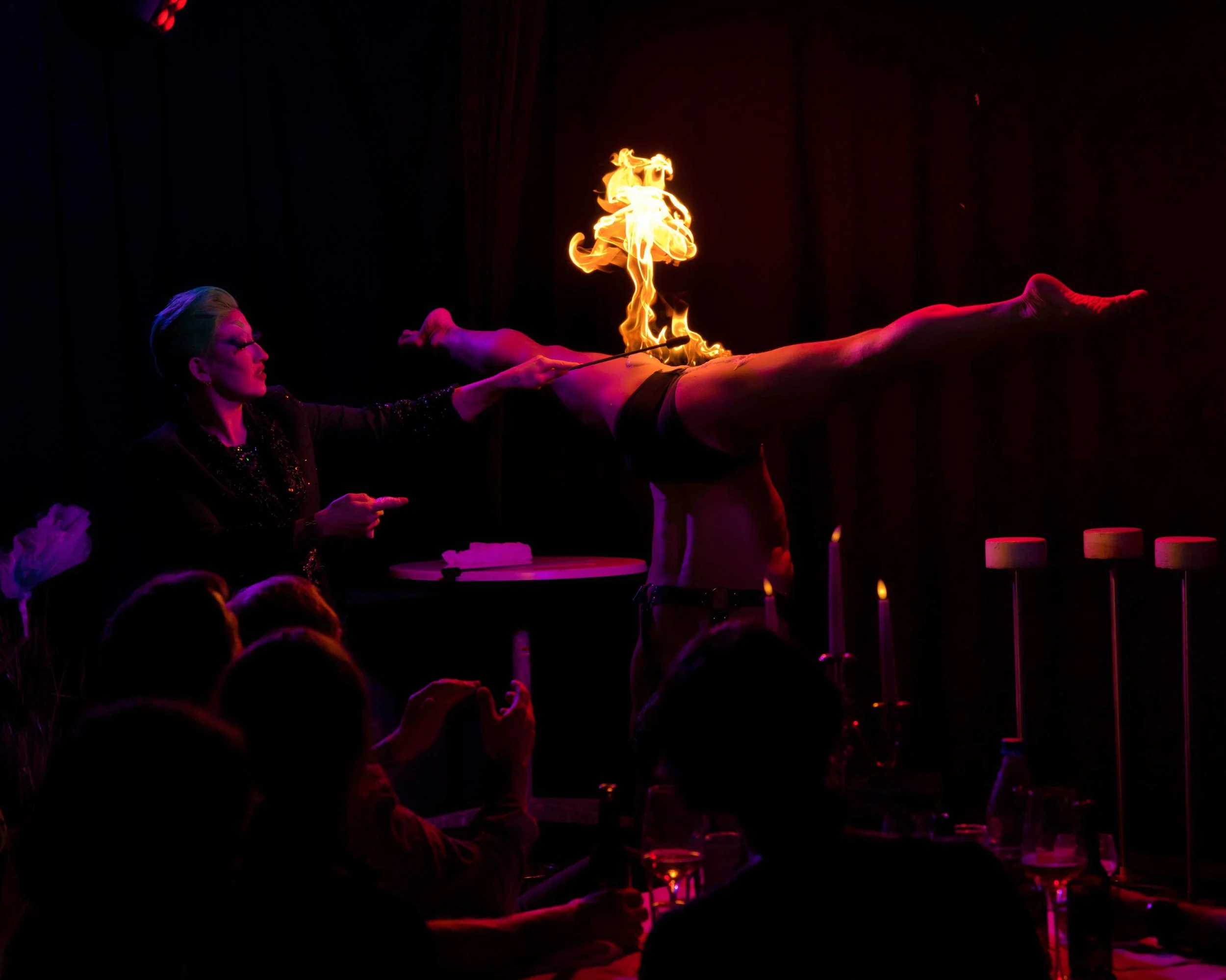 Feuershow bei einer Zirkusperformance mit einer Artistin, die auf einem Tisch balanciert und von einer Frau angeleitet wird, während Zuschauer zuschauen.