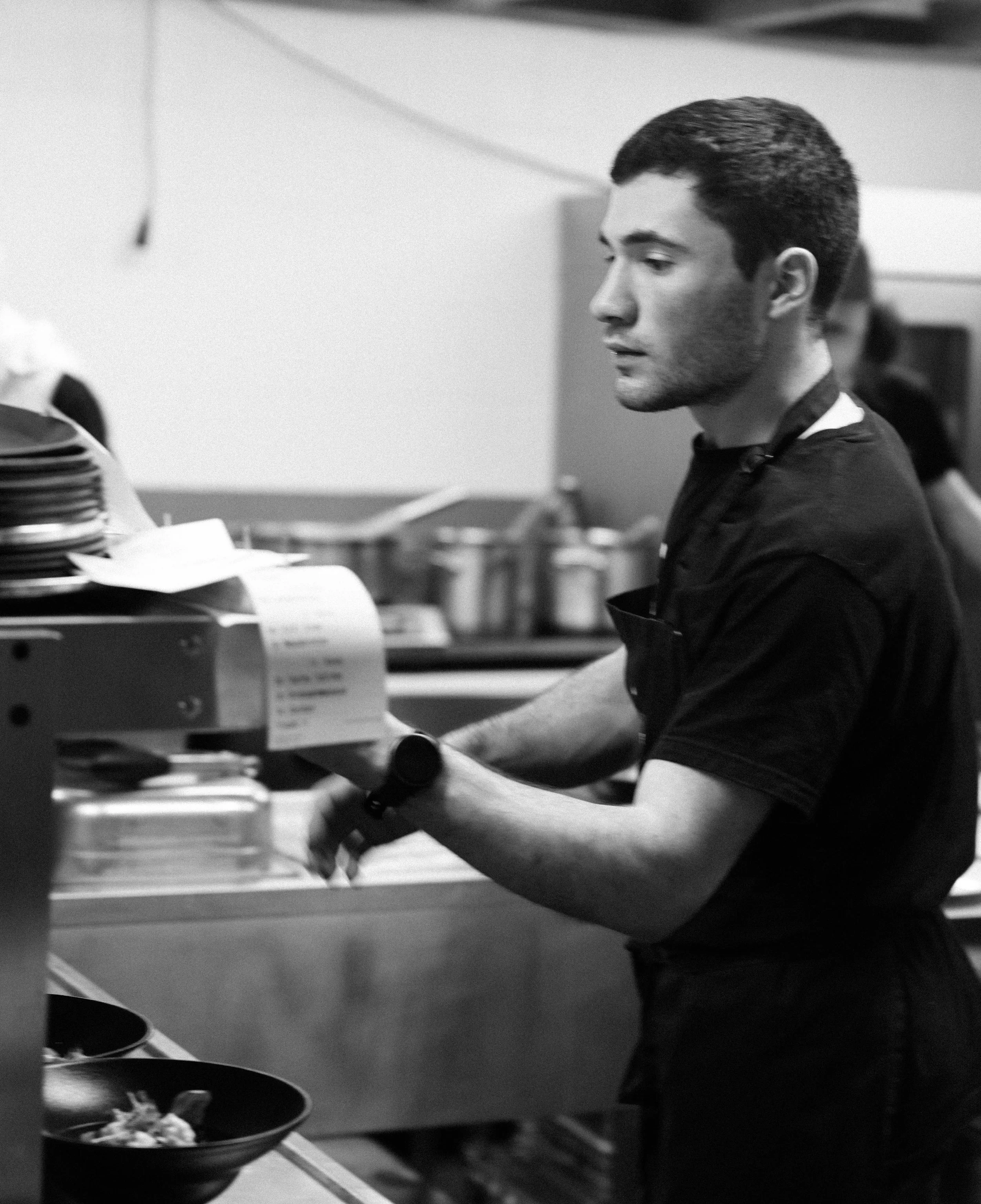 Ein junger Mann in Schwarz-Weiß steht in einer Küche, scheint bei der Arbeit zu sein, und trägt eine schwarze Uniform mit einem schwarzen Armband oder Uhr am Handgelenk, während er neben einem Küchenbuffet steht.
