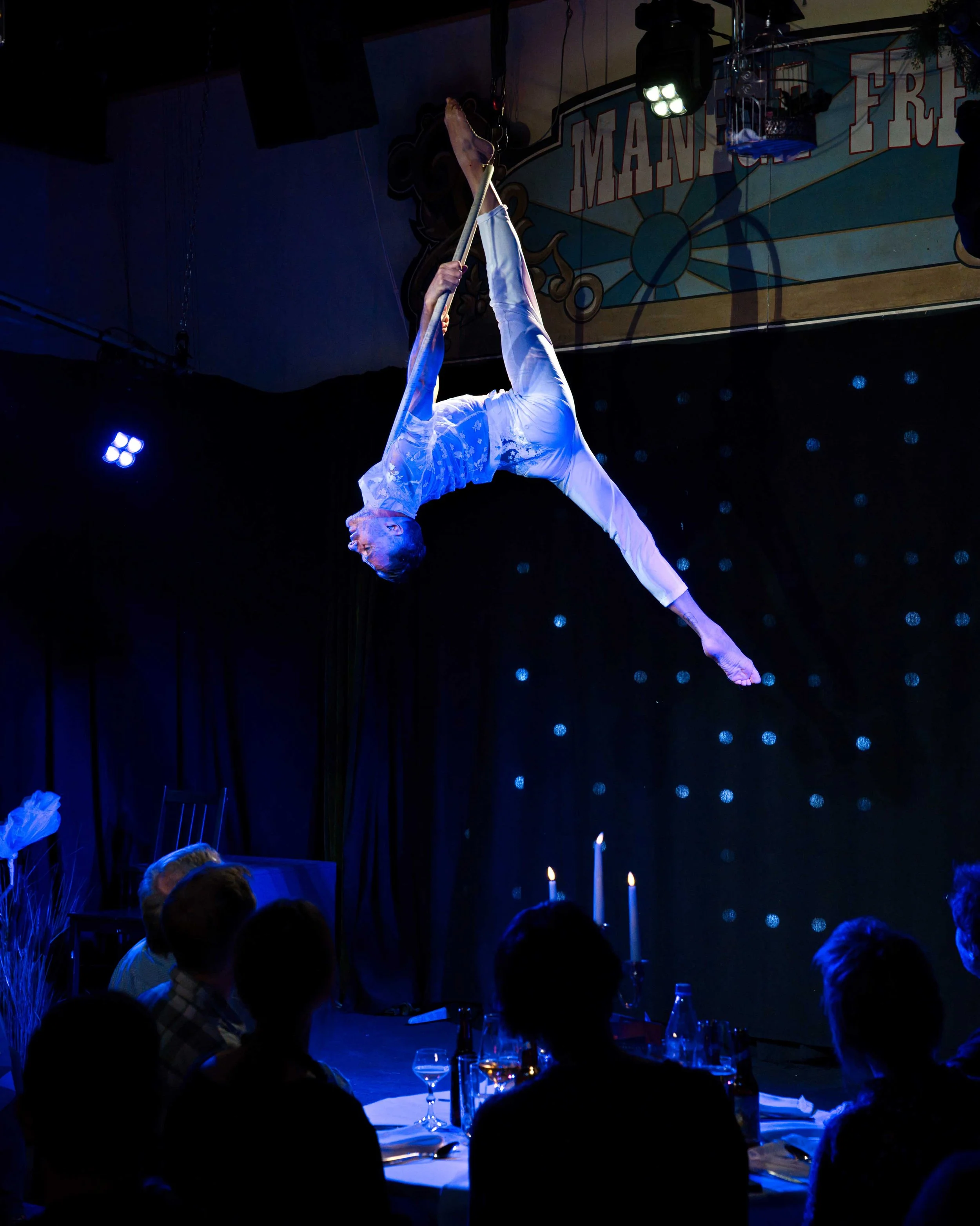 Ein Akrobat hängt an einem Seil über einem Tisch mit Gästen, während er eine Vorstellung im Theater macht.