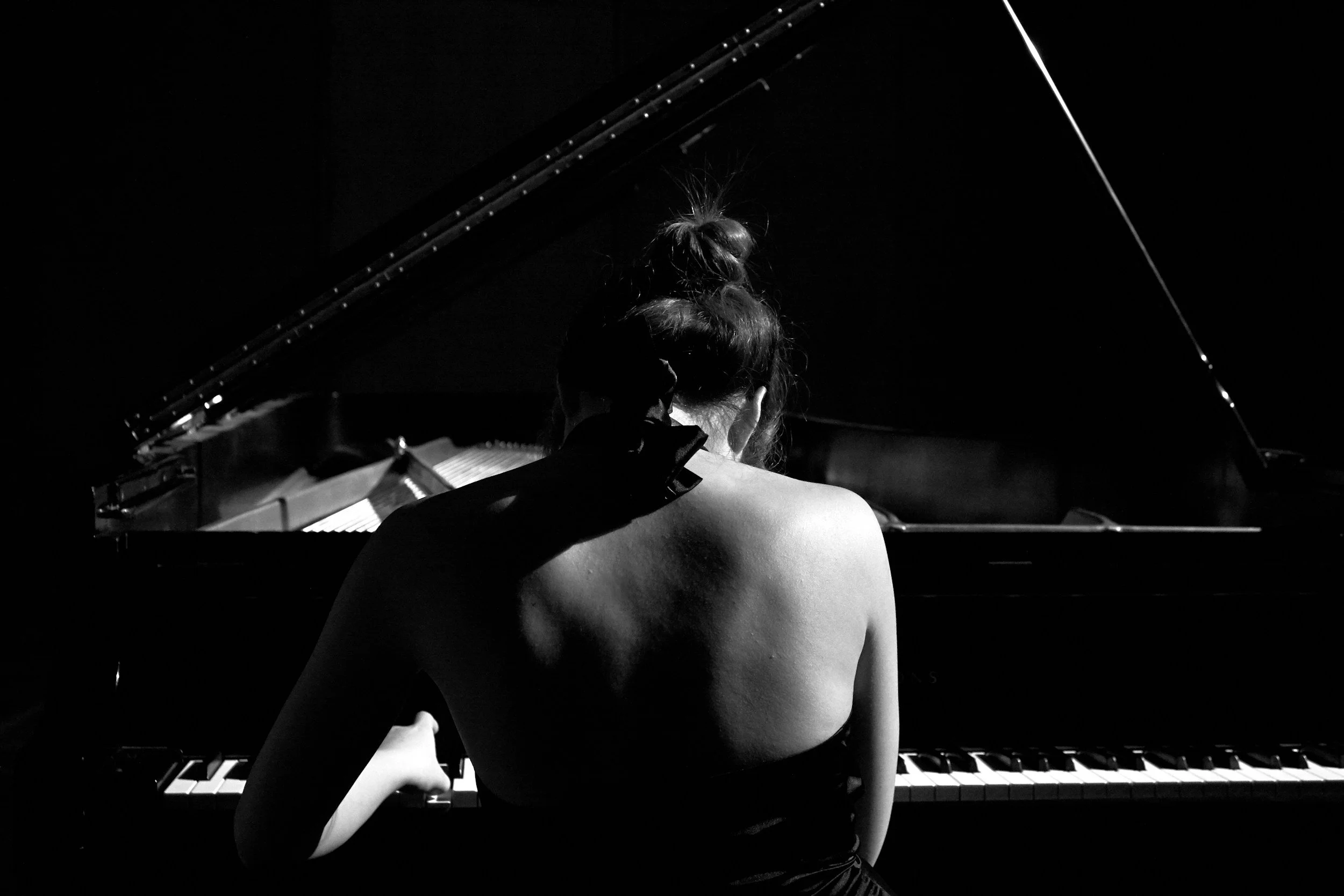 A black-and-white photo of a woman with her back to the camera, sitting at a grand piano, with her hair tied up in a messy bun. Ambient light highlights her shoulder, back, and the piano keys.