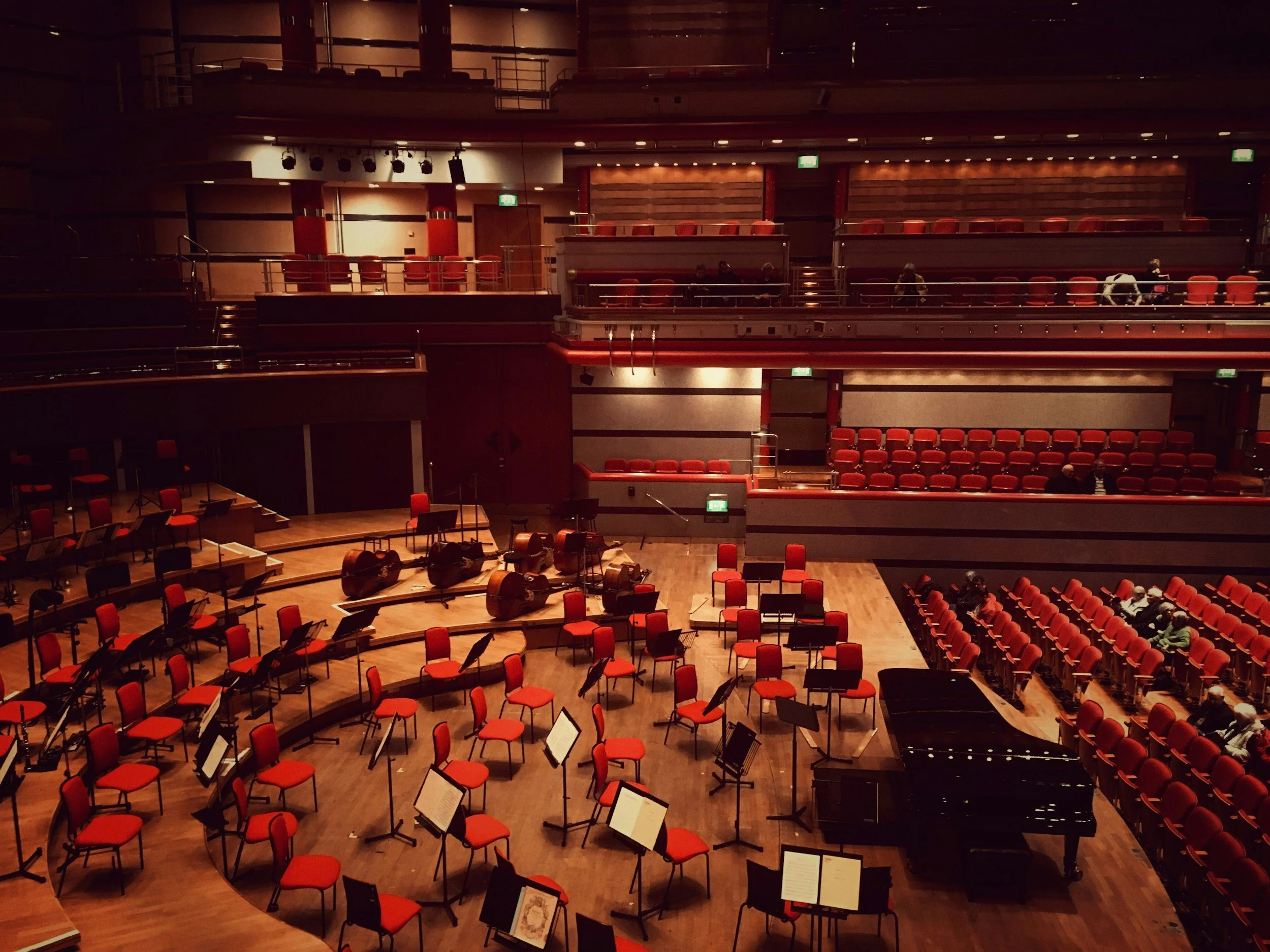 The image shows an empty concert hall with tiered seating, red chairs, and scattered music stands and musical instruments on the stage, with a few people seated in the audience area.