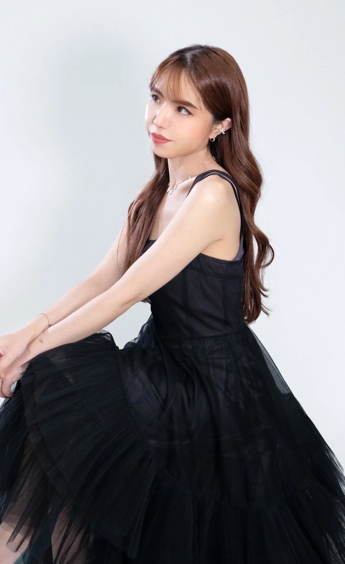 Young woman with long brown hair in a black dress, sitting with legs crossed against a plain white background.