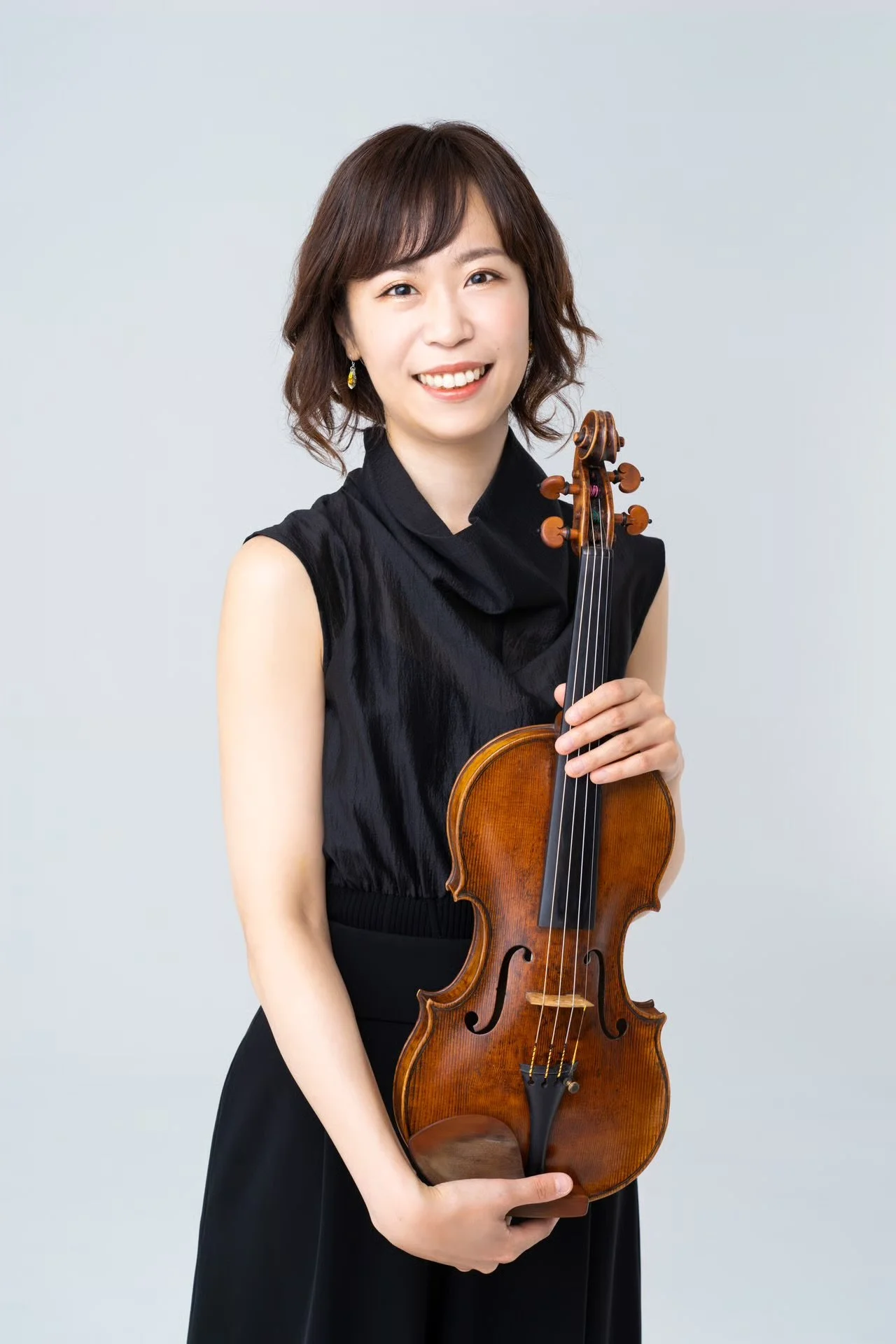 A woman with short brown hair holding a violin and smiling at the camera, wearing a black sleeveless top and earrings, against a light gray background.