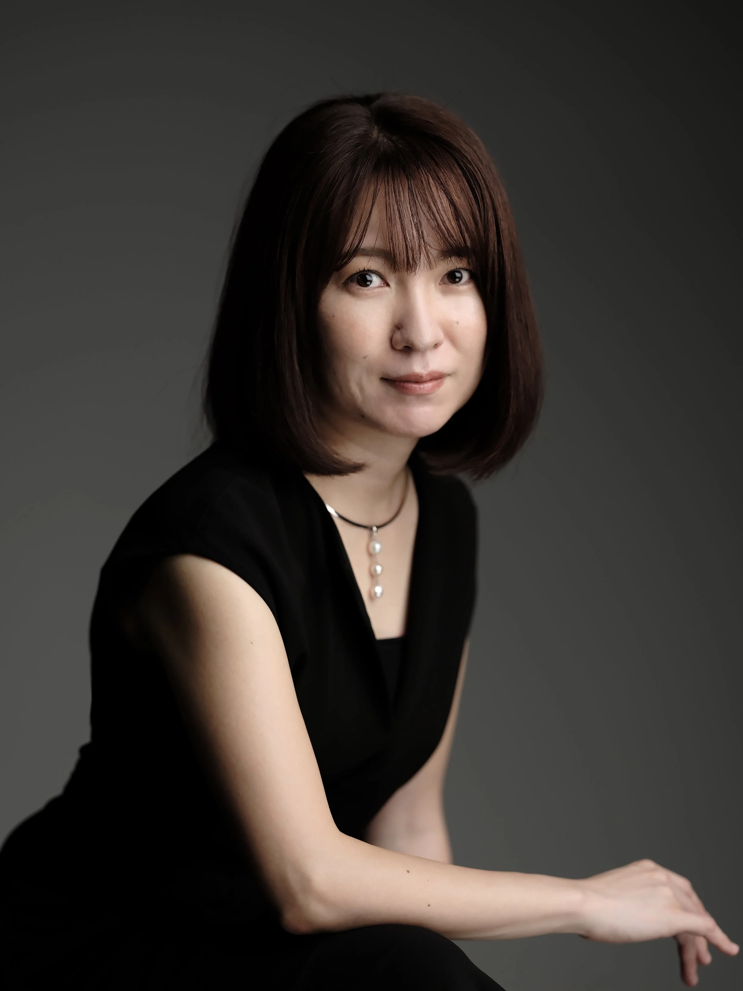 A woman with shoulder-length dark brown hair, wearing a black top and a pearl necklace, is sitting against a dark gray background. She is looking at the camera with a slight smile, and her right hand rests on her lap.