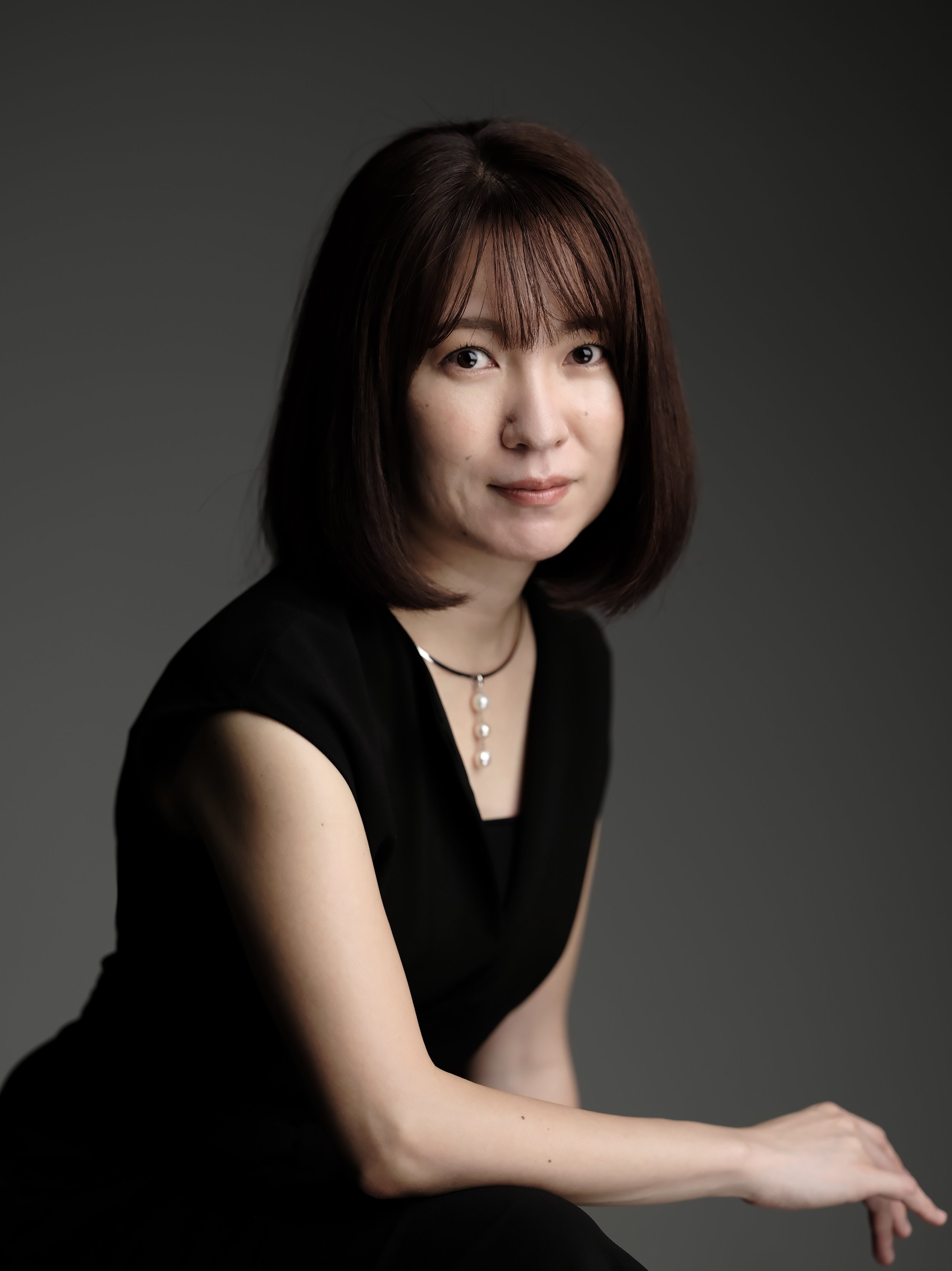 Portrait of a woman with shoulder-length brown hair, wearing a black top and a pearl necklace, sitting against a dark gray background.