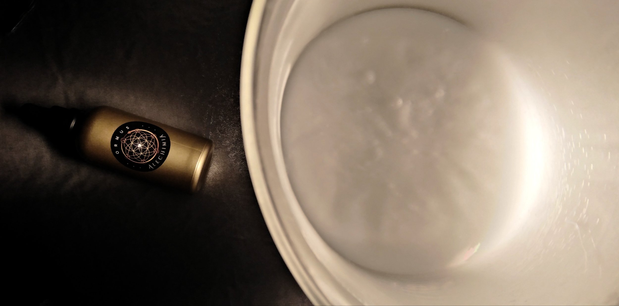 A metallic gold spray bottle labeled 'Ormus Alechimia' lying on a black surface next to an empty metal bucket or container.