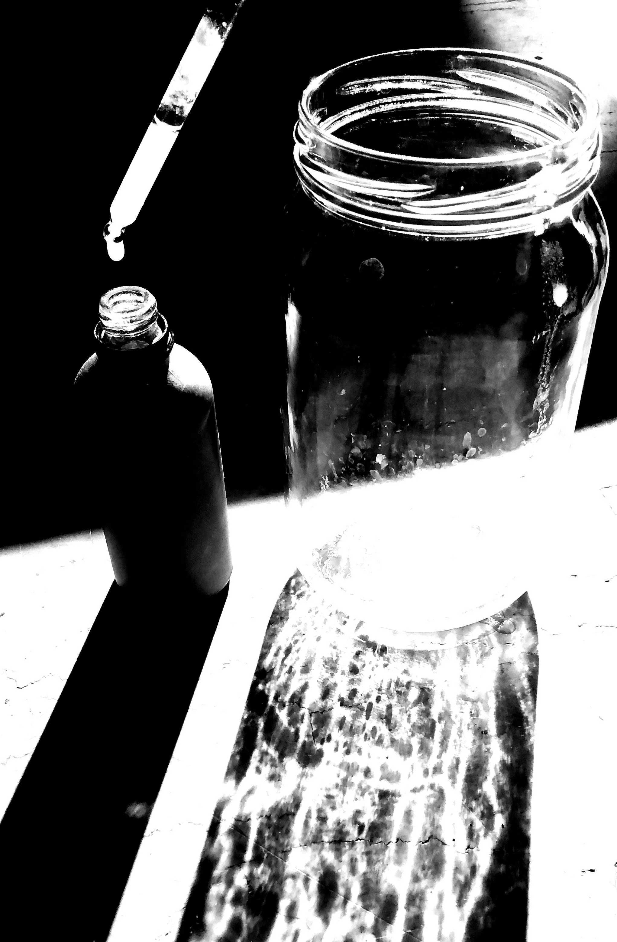 A black and white photo showing a dropper above an open glass jar and a smaller bottle on a surface, with sunlight creating reflections and shadows.