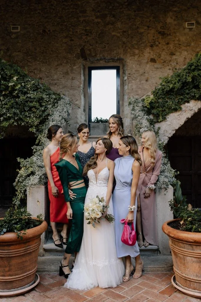 A bride in a white wedding dress holding a bouquet gathers with six women in colorful dresses on a stone staircase inside a rustic building with stone walls and greenery.