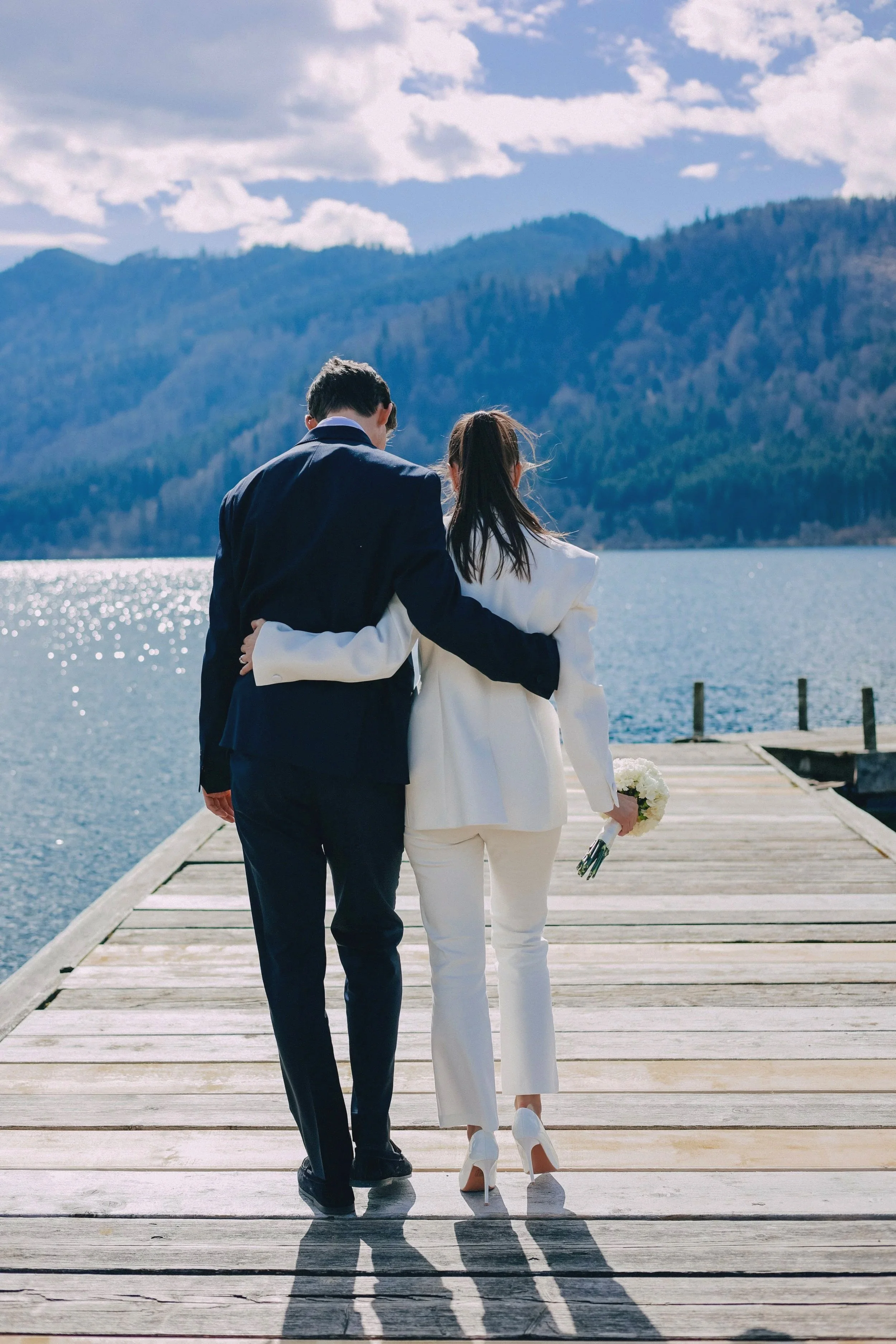A couple dressed in formal white and navy suits walking arm-in-arm on a wooden dock by a lake with mountain scenery in the background.