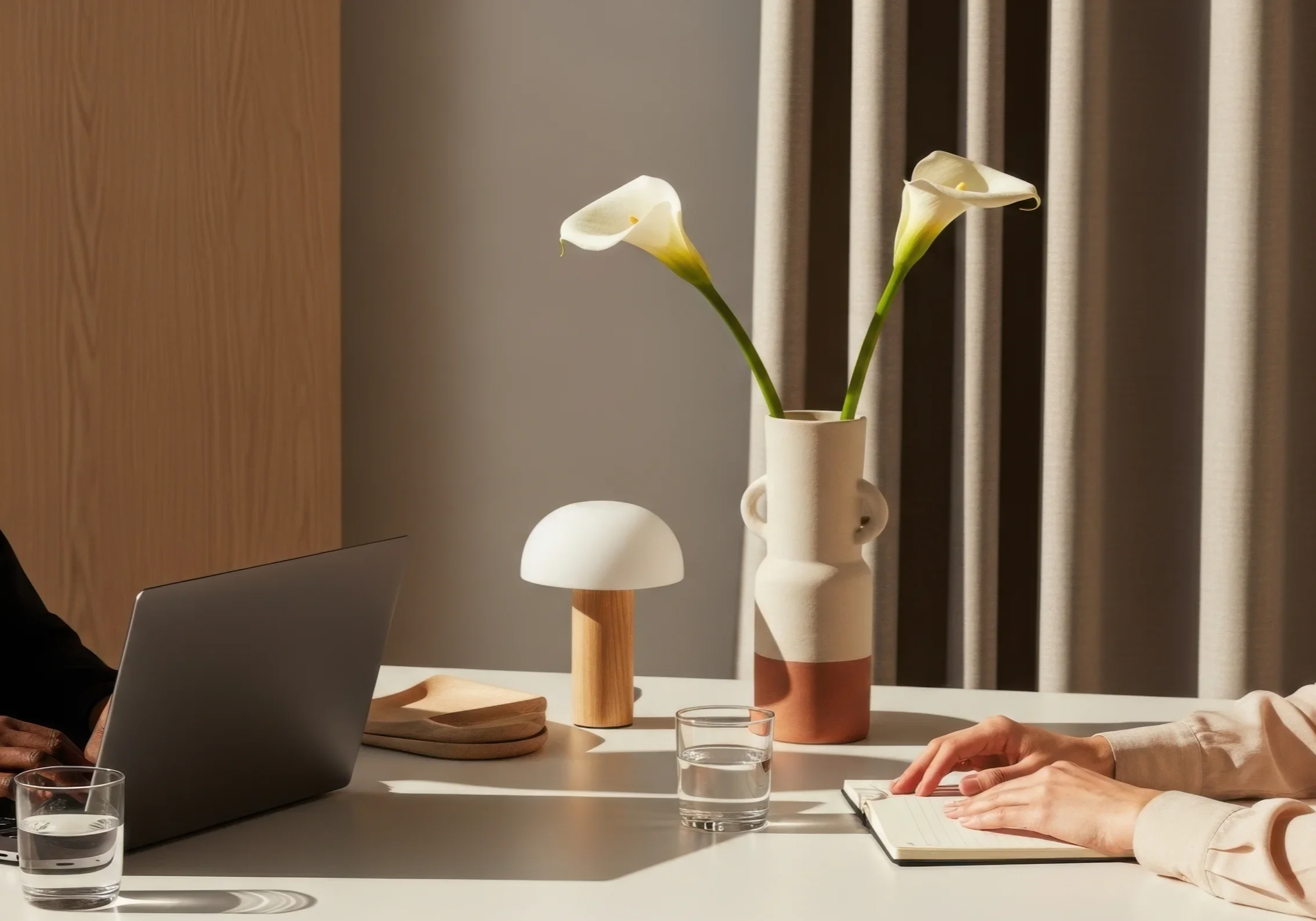 Decorative table setting with a white vase holding two calla lilies, a small mushroom-shaped lamp, a glass of water, a notebook, and two glasses of water, with parts of two people visible.