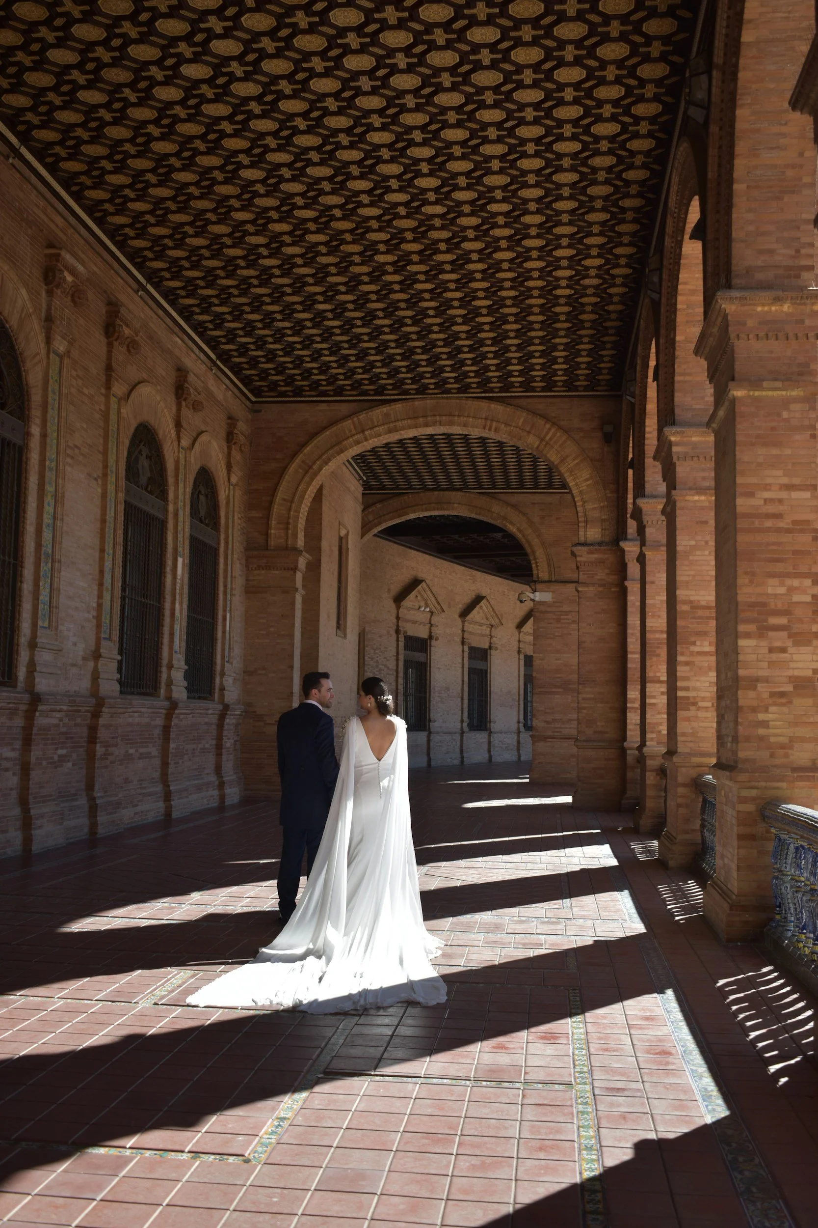 A bride and groom standing together on a tiled walkway under an ornate, arched covered walkway with brick walls and decorative ceiling patterns.
