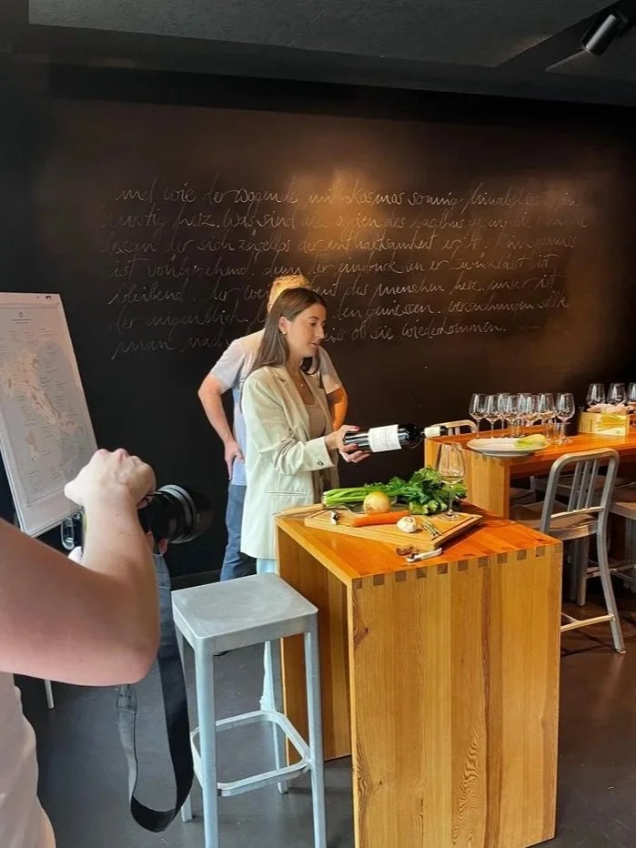A woman in a beige blazer holds a wine bottle while standing at a wooden table with vegetables, including celery, onions, and garlic, in a restaurant or wine tasting room. There are empty wine glasses on a counter behind her, and a person is taking a