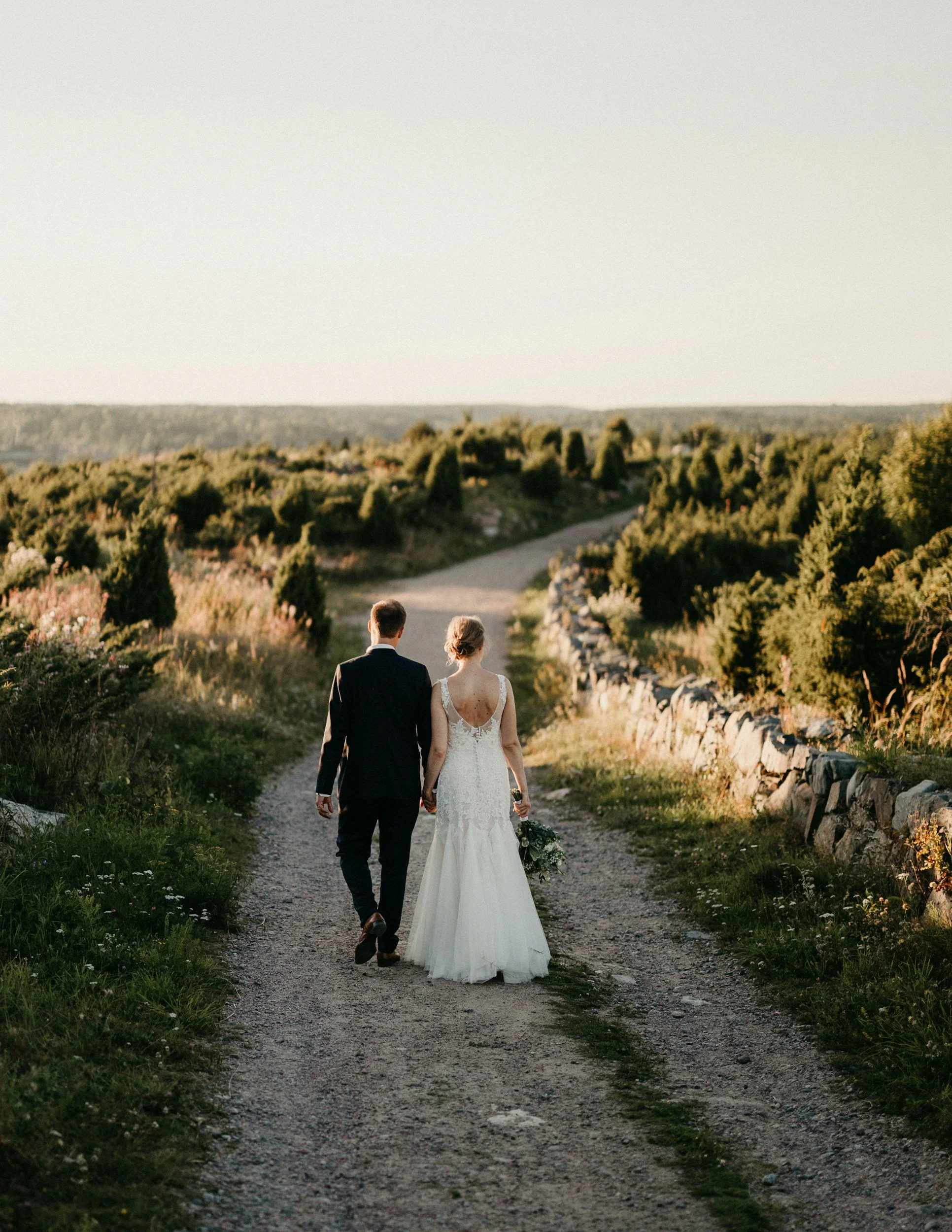 A newlywed couple walking hand in hand on a gravel path in a scenic outdoor setting during sunset.