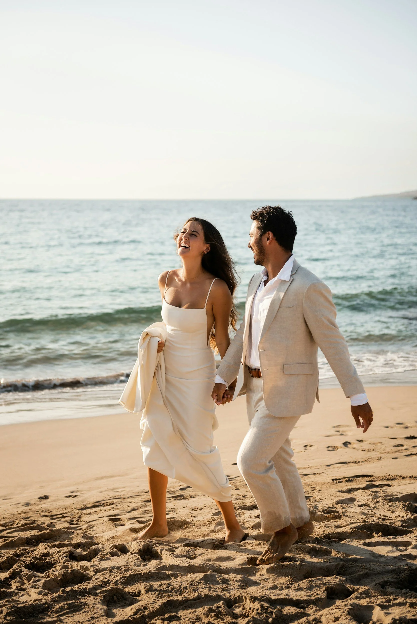 A happy couple in wedding attire walking hand in hand on the beach near the ocean.