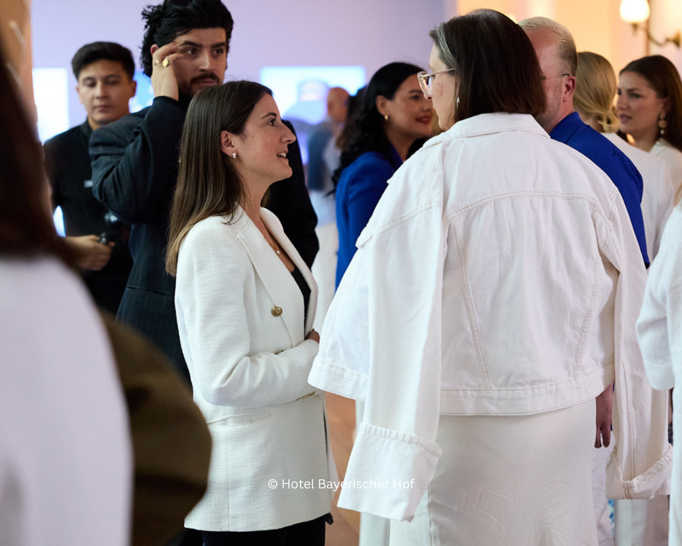 Group of people engaged in conversation at a professional event, with women in white and blue and women in white dresses.